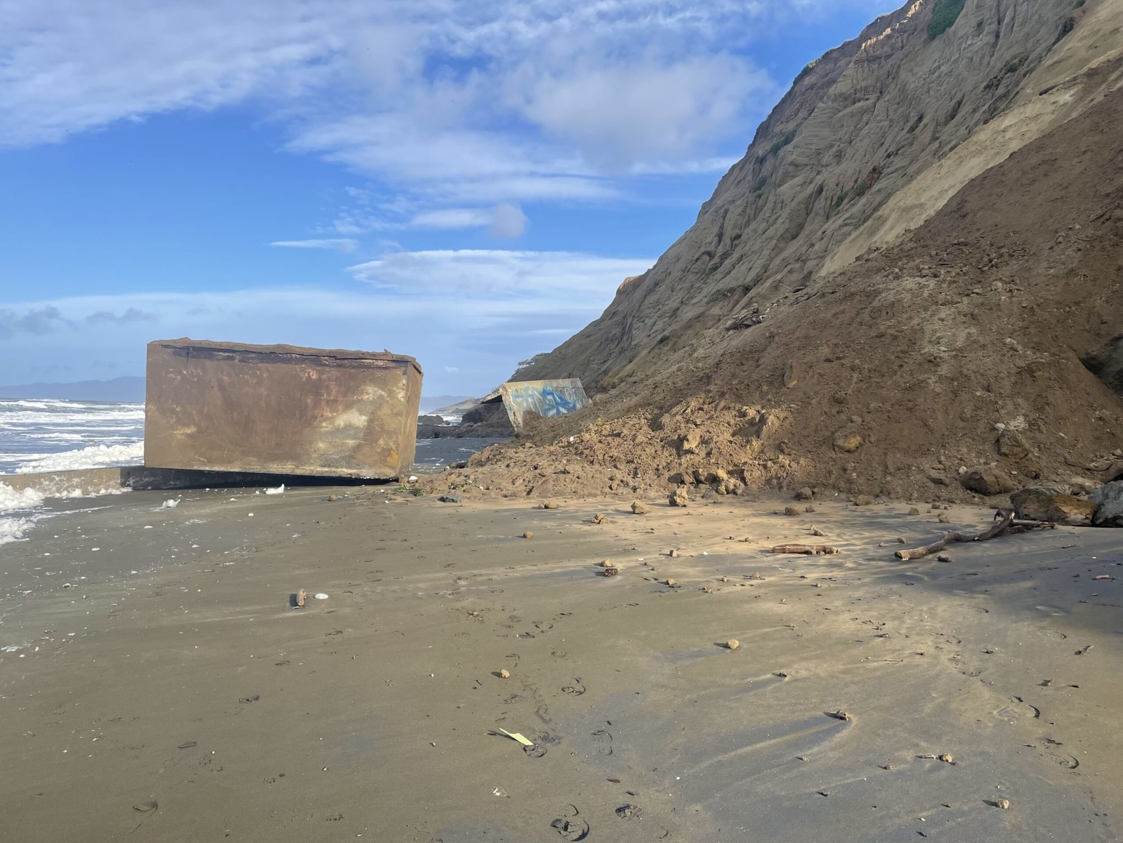 A sandy beach with large, rusted concrete blocks near a steep eroded cliff and scattered debris under a partly cloudy blue sky.
