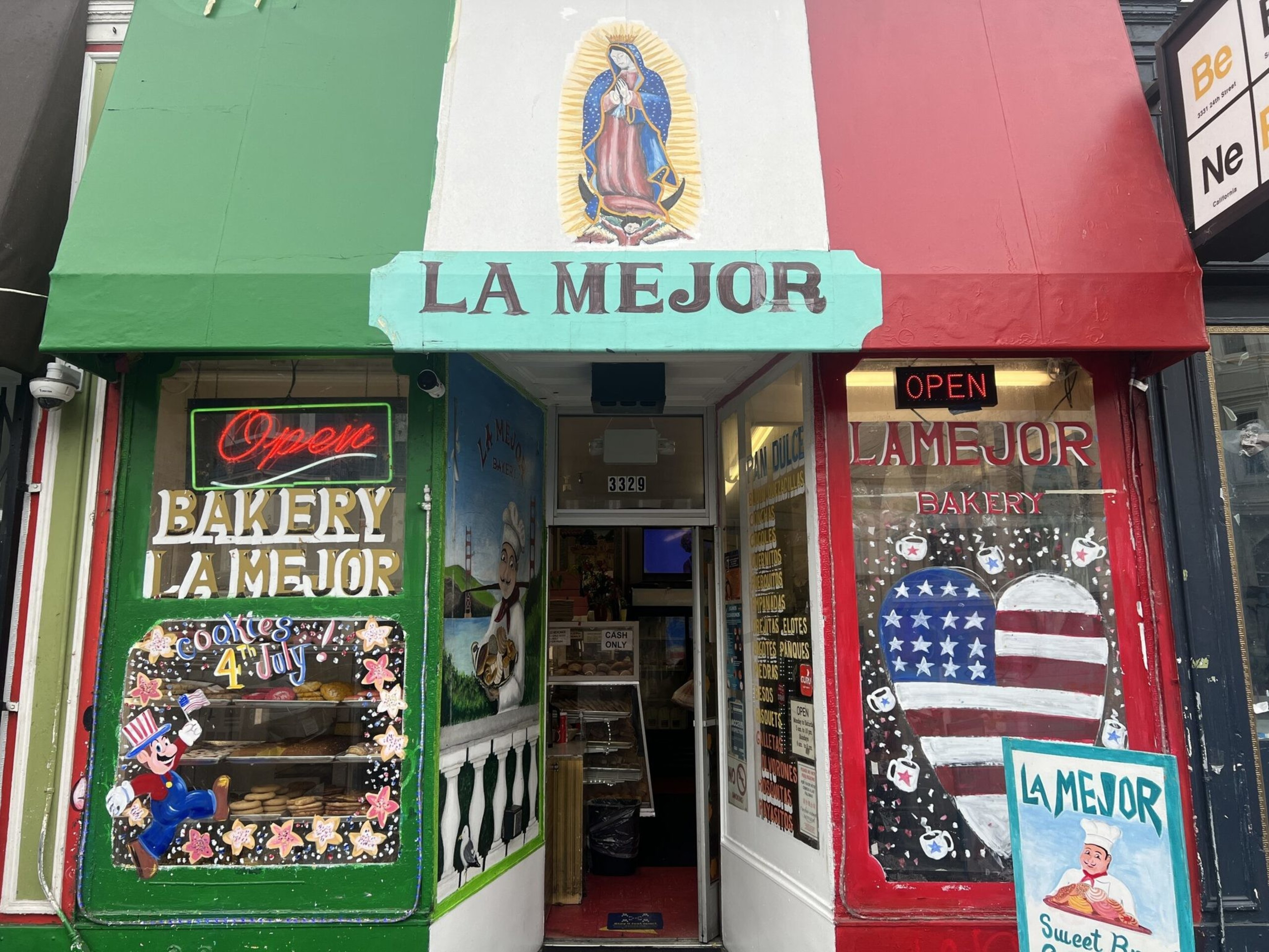 A bakery storefront painted in green, white, and red with "LA MEJOR" signs, patriotic 4th of July cookie decorations, and "Open" neon signs.