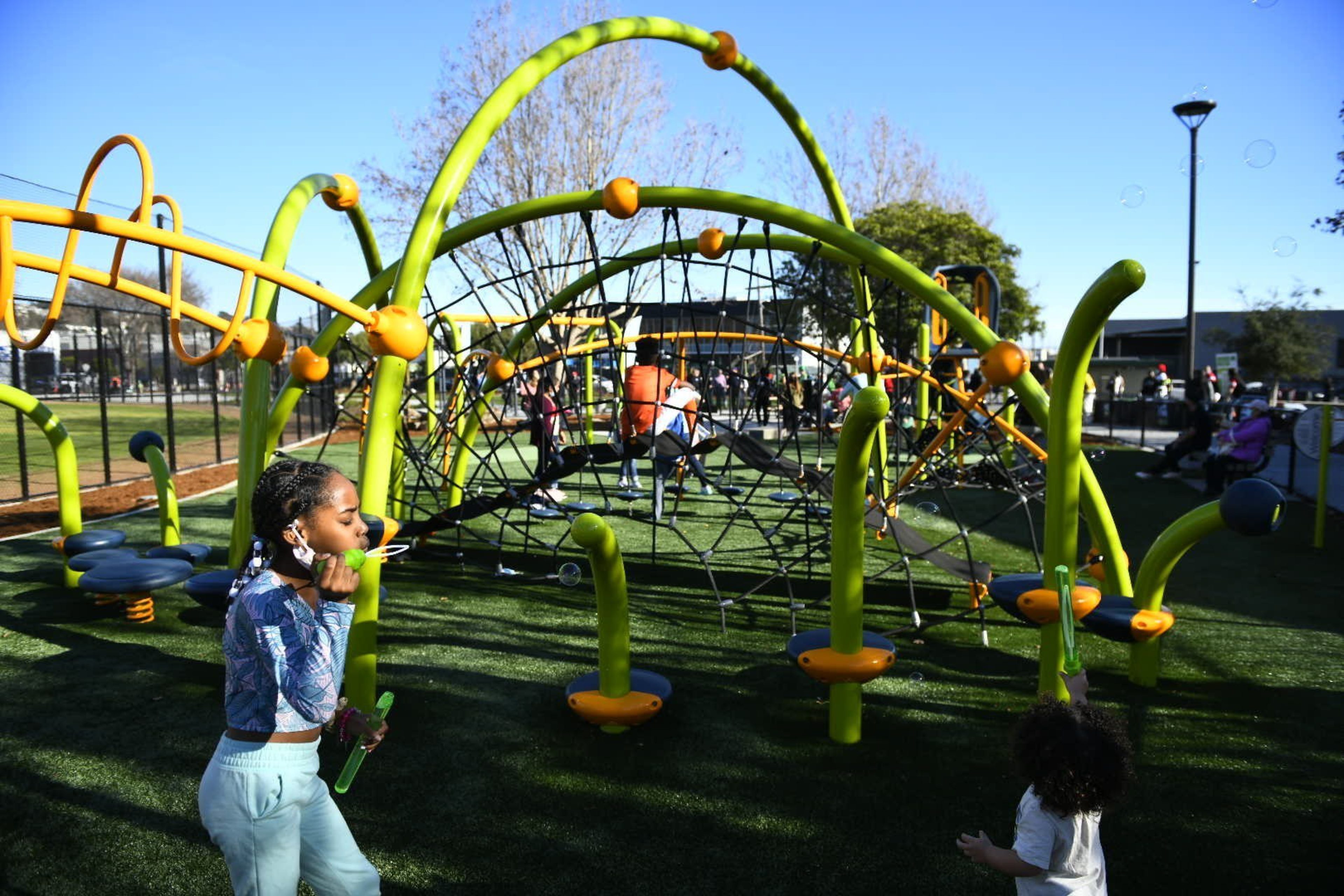 Children play on a colorful green and orange playground structure with ropes and slides under a clear blue sky.