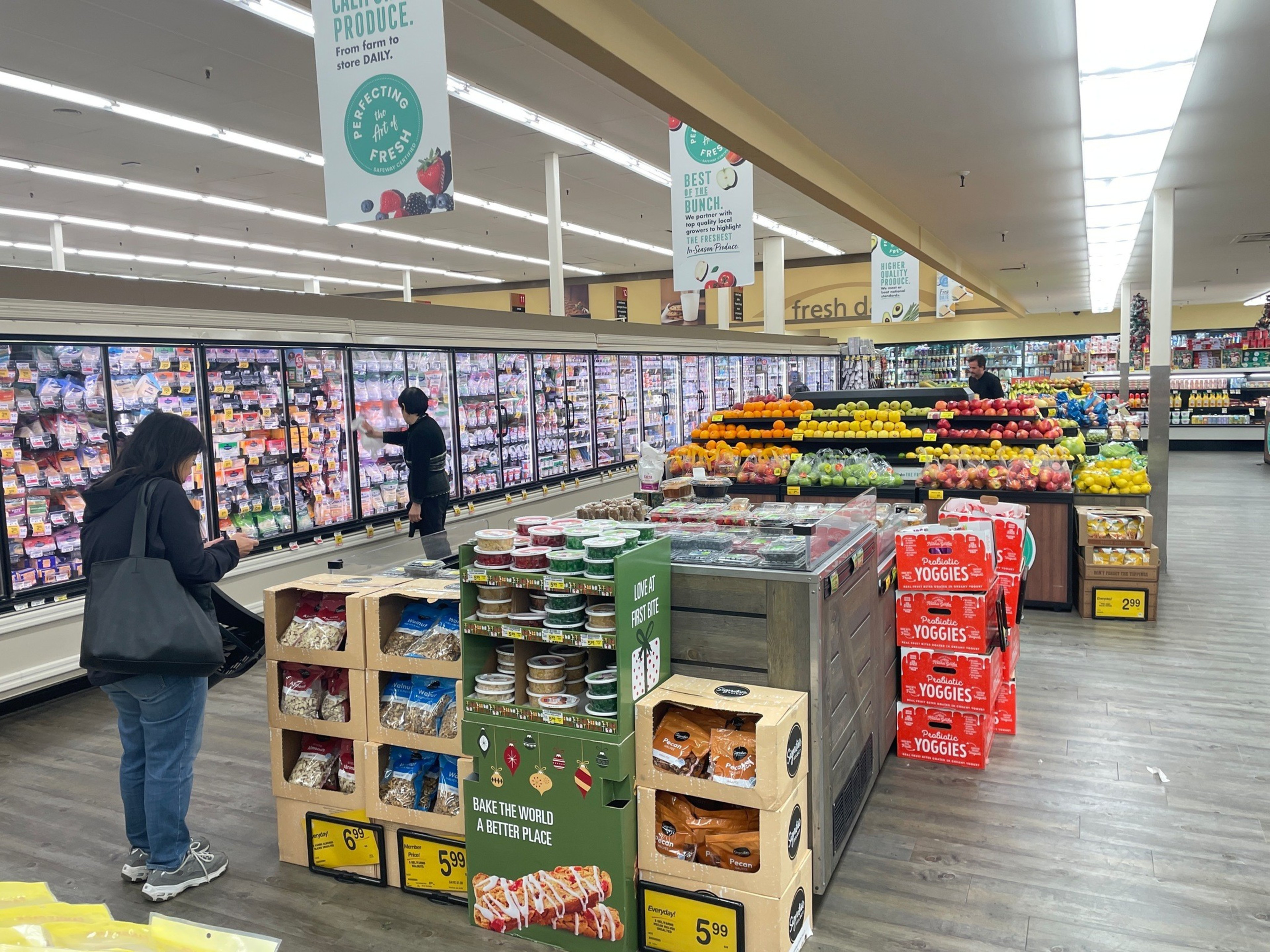 A grocery store aisle with refrigerated meat cases along the left, fresh fruit displays in the center, and shoppers browsing.