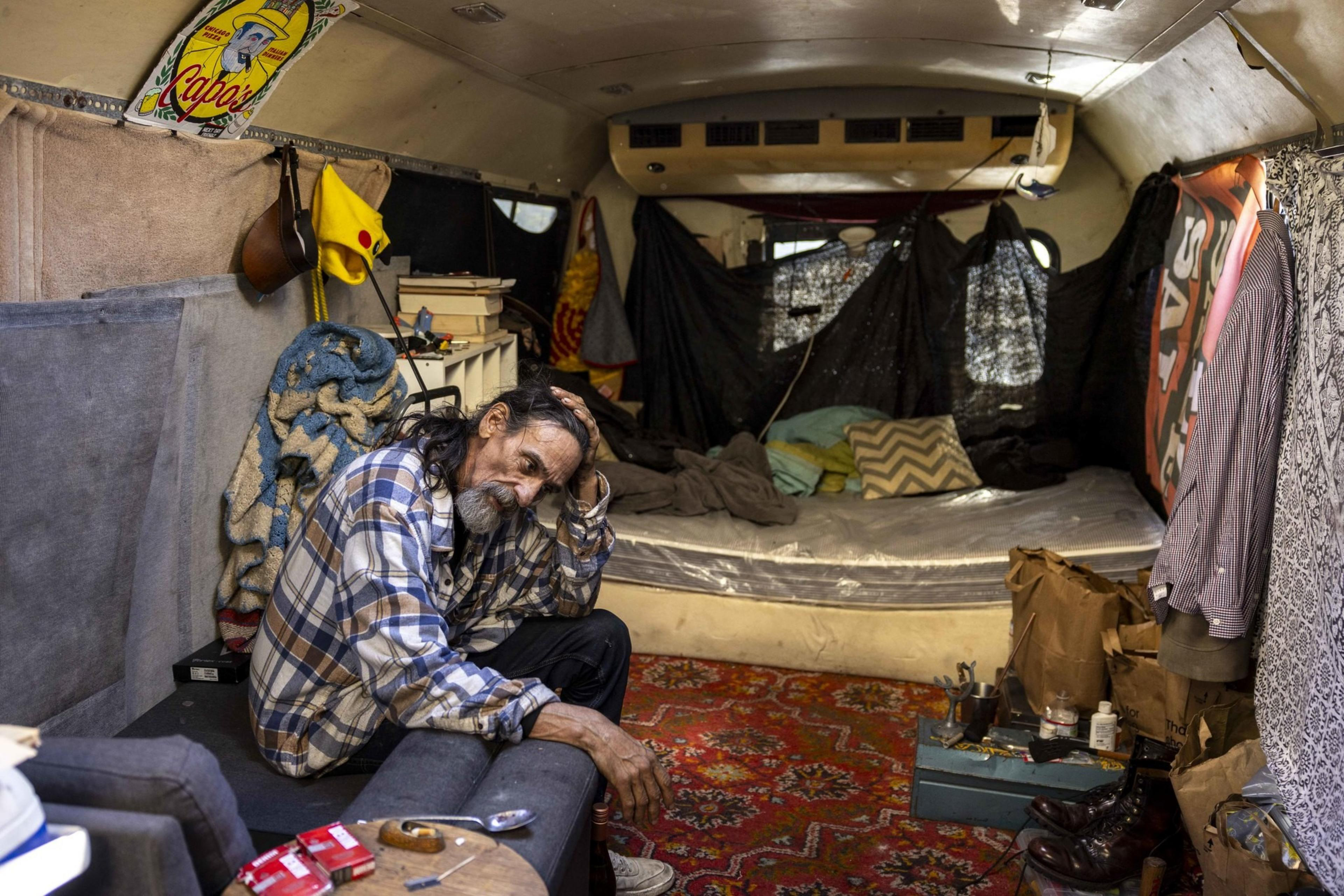 A man in a plaid shirt sits on a couch inside a cluttered trailer. The space features a sleeping area with pillows, various items scattered around, and a vibrant rug.