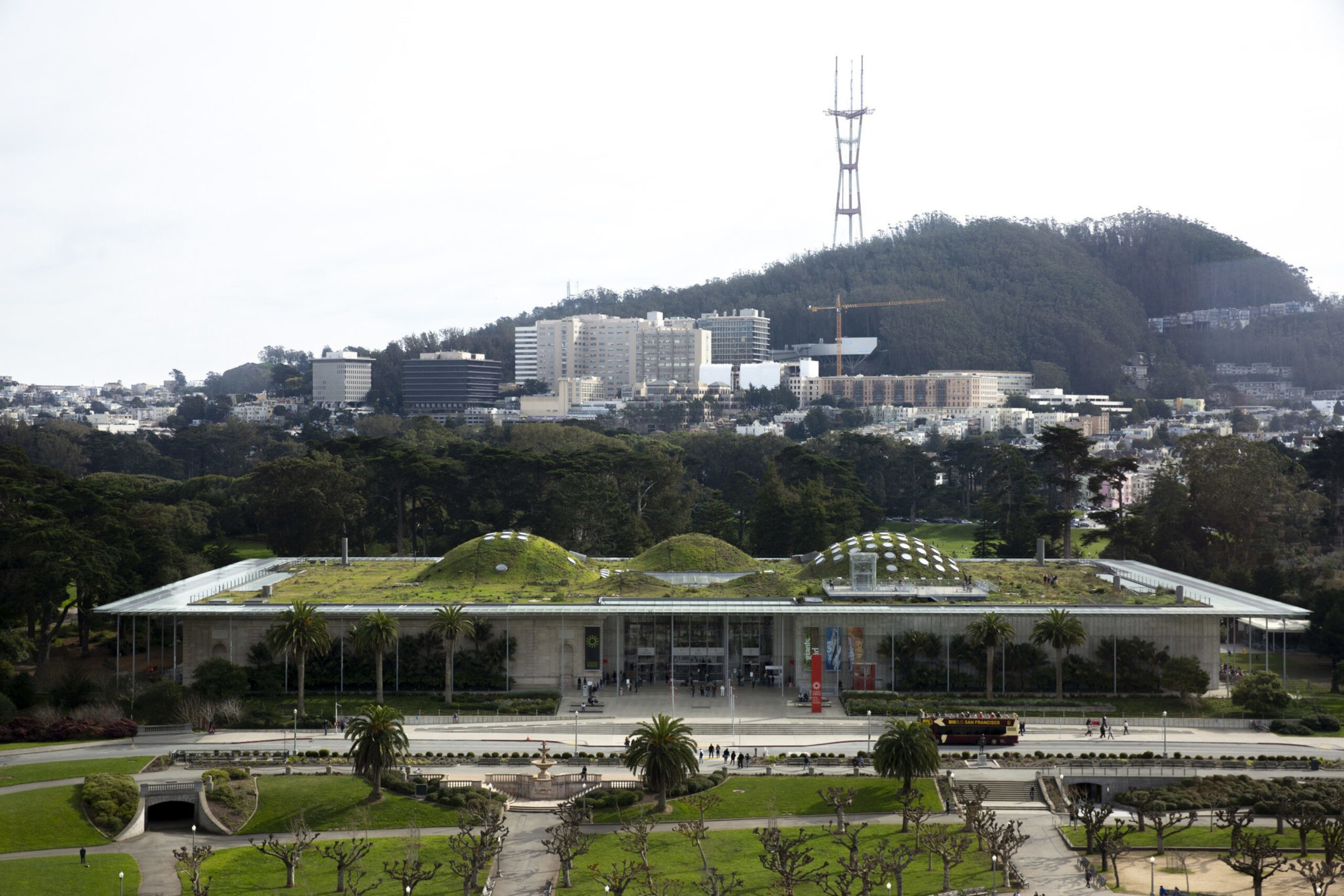A large modern building with a green, mound-covered roof sits in front of a forested hill topped by a tall communications tower.