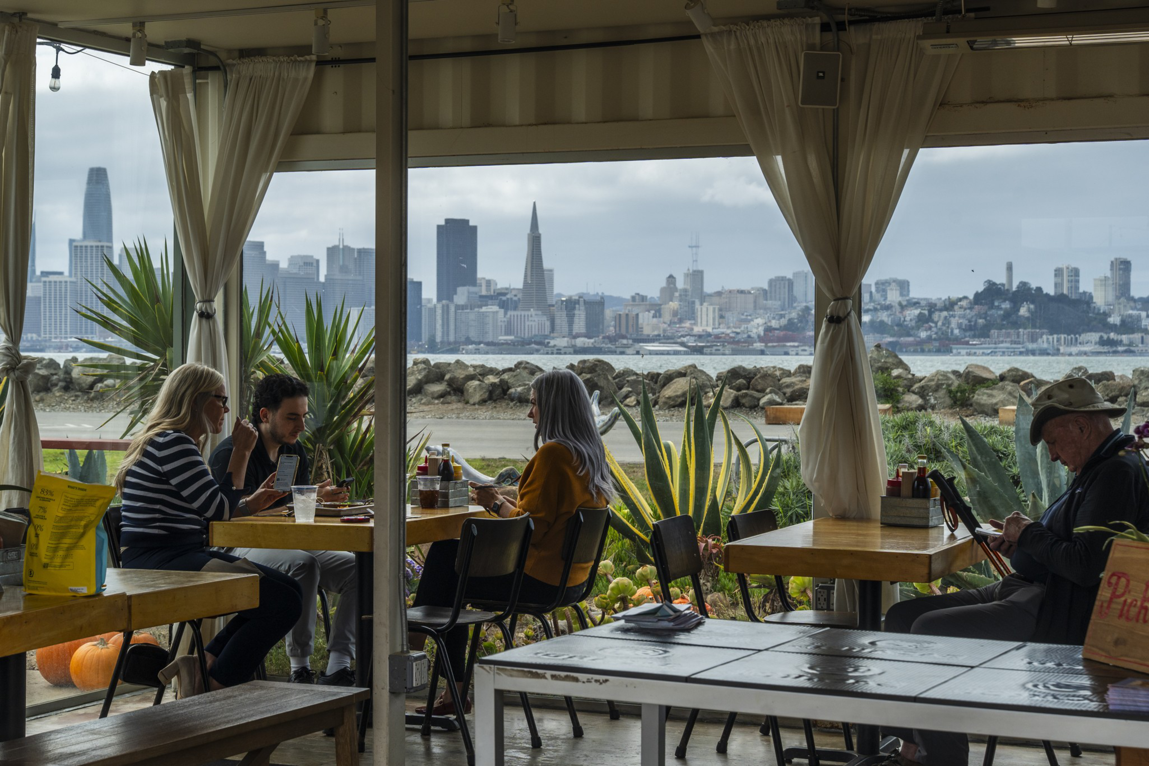 Three people sit at a table near a window with a view of a city skyline across water, while an older man sits alone at another table using a device.
