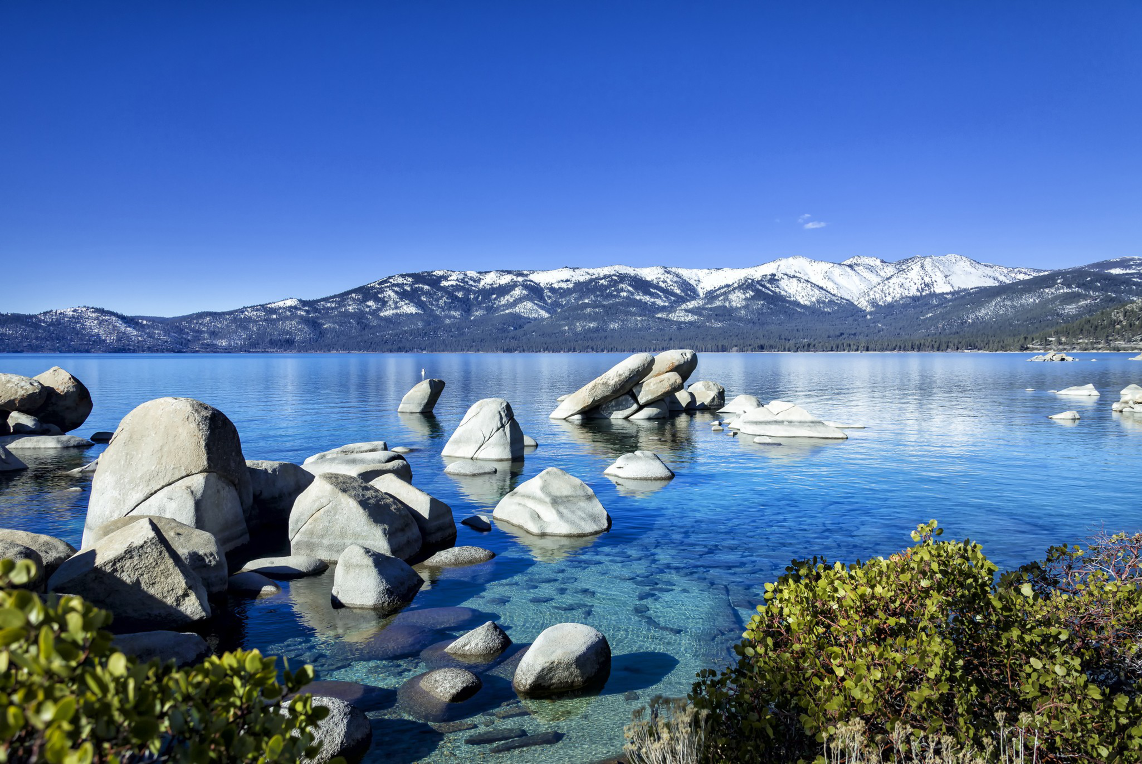 Clear blue lake water surrounds large smooth rocks with snow-capped mountains and a bright blue sky in the background, framed by green bushes in the foreground.