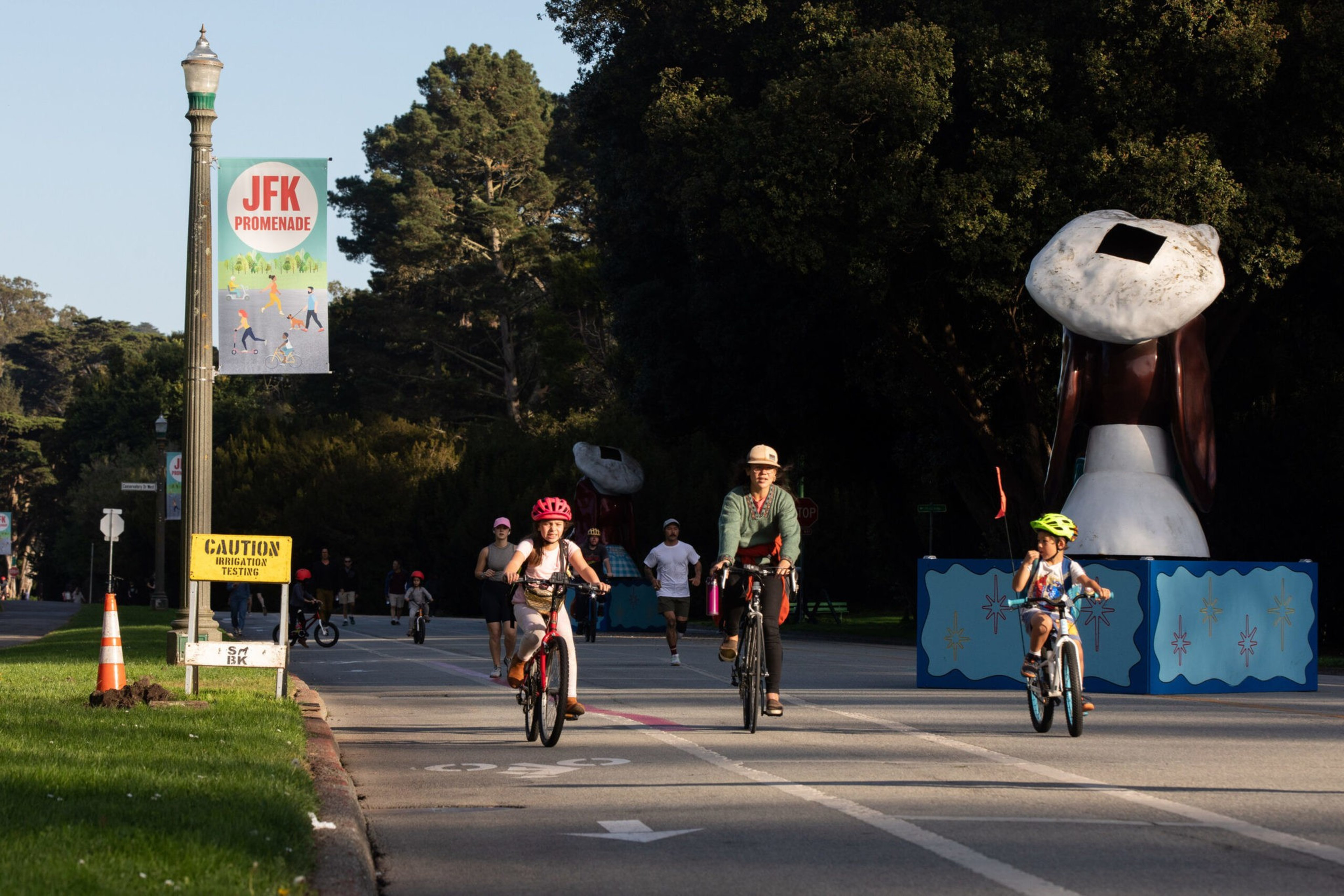 Children and an adult ride bikes on a road lined with trees, a lamp post with a "JFK Promenade" banner, and a large Snoopy statue nearby.