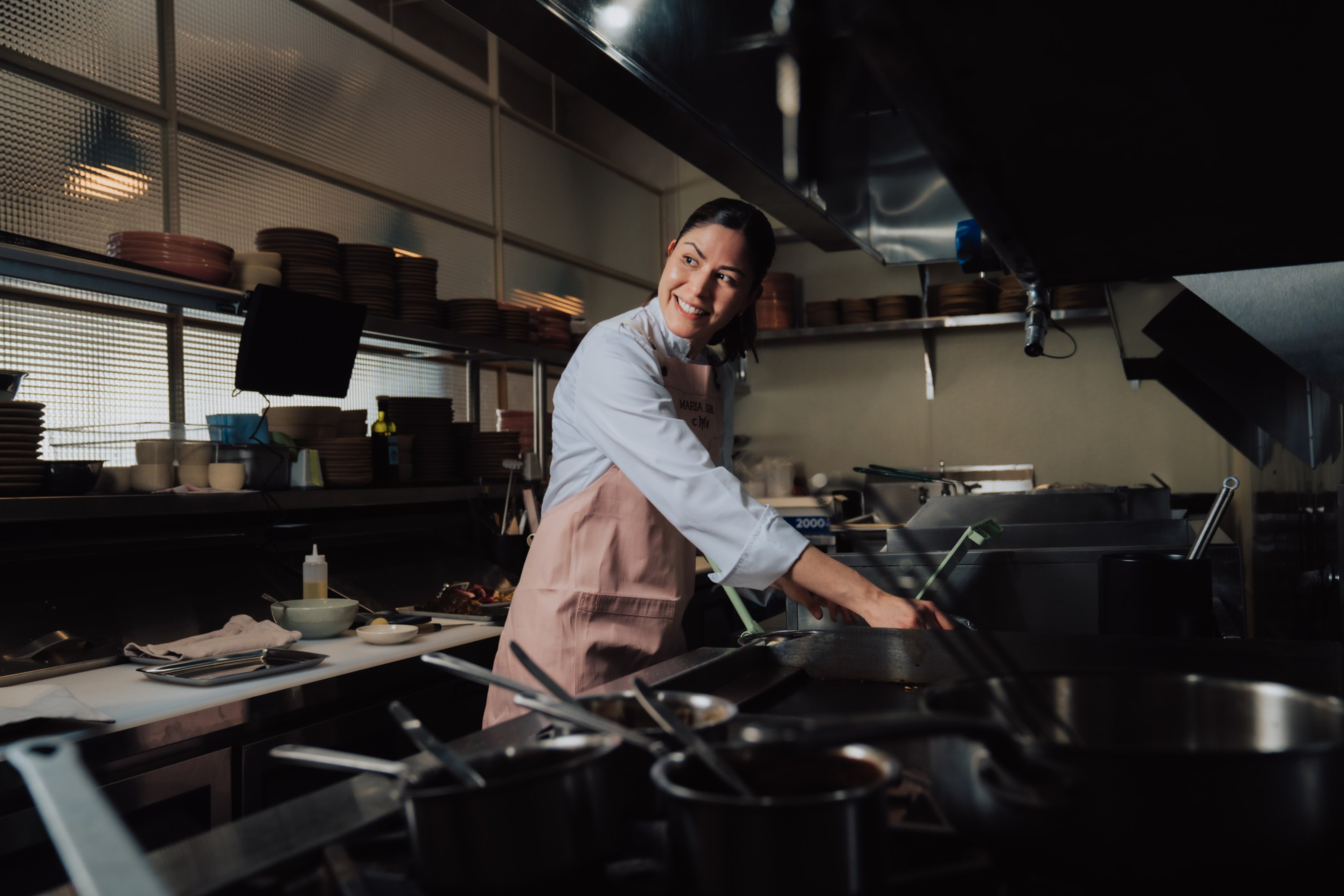 A smiling chef in a white shirt and pink apron cooks on a stove in a dimly lit commercial kitchen filled with pots, pans, and dishes.
