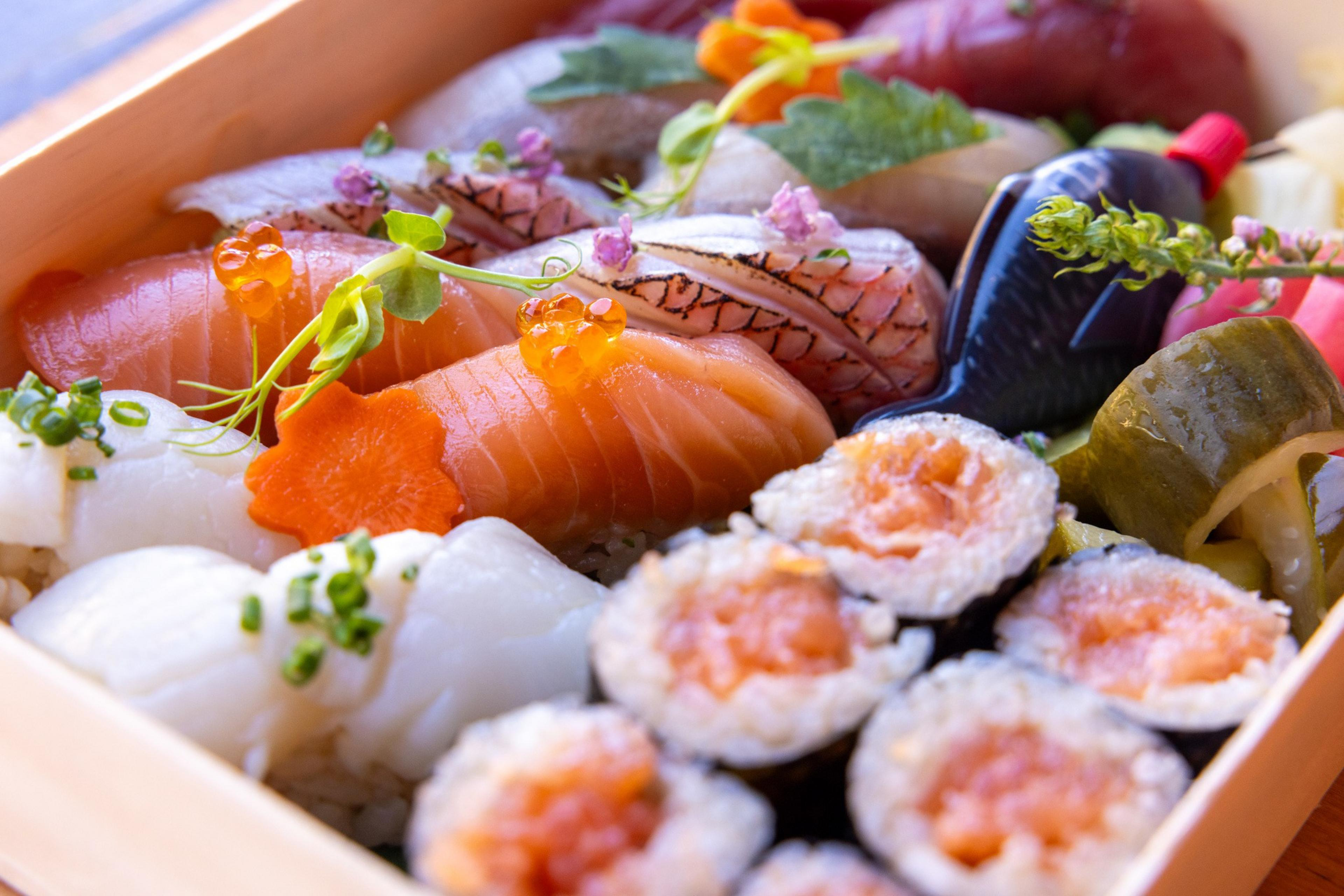 The image shows a wooden box filled with various sushi pieces, including nigiri and maki rolls. Garnishes and a small bottle of soy sauce are also visible.