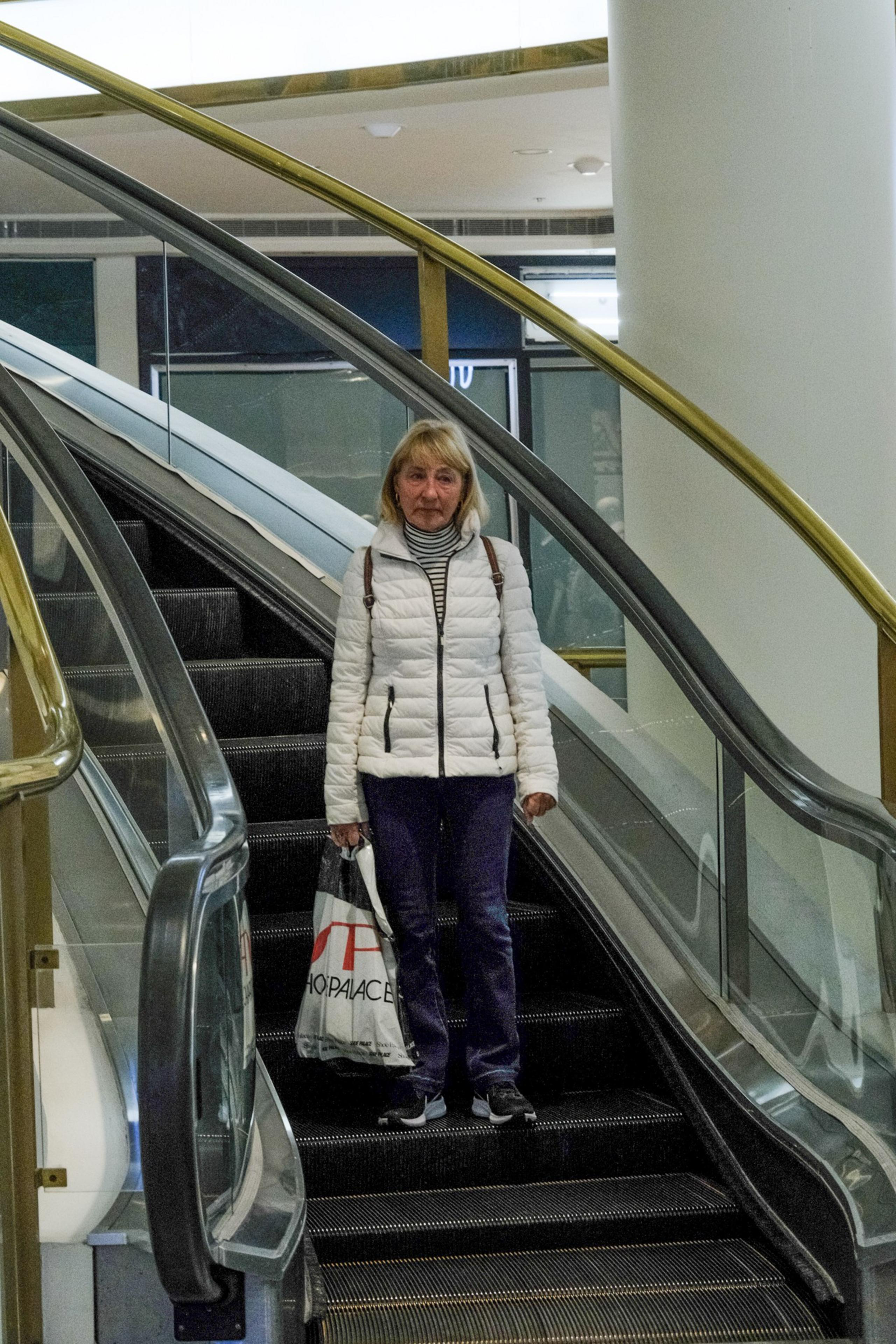 A person descends an escalator holding a shopping bag, wearing a white jacket, striped shirt, and blue pants.
