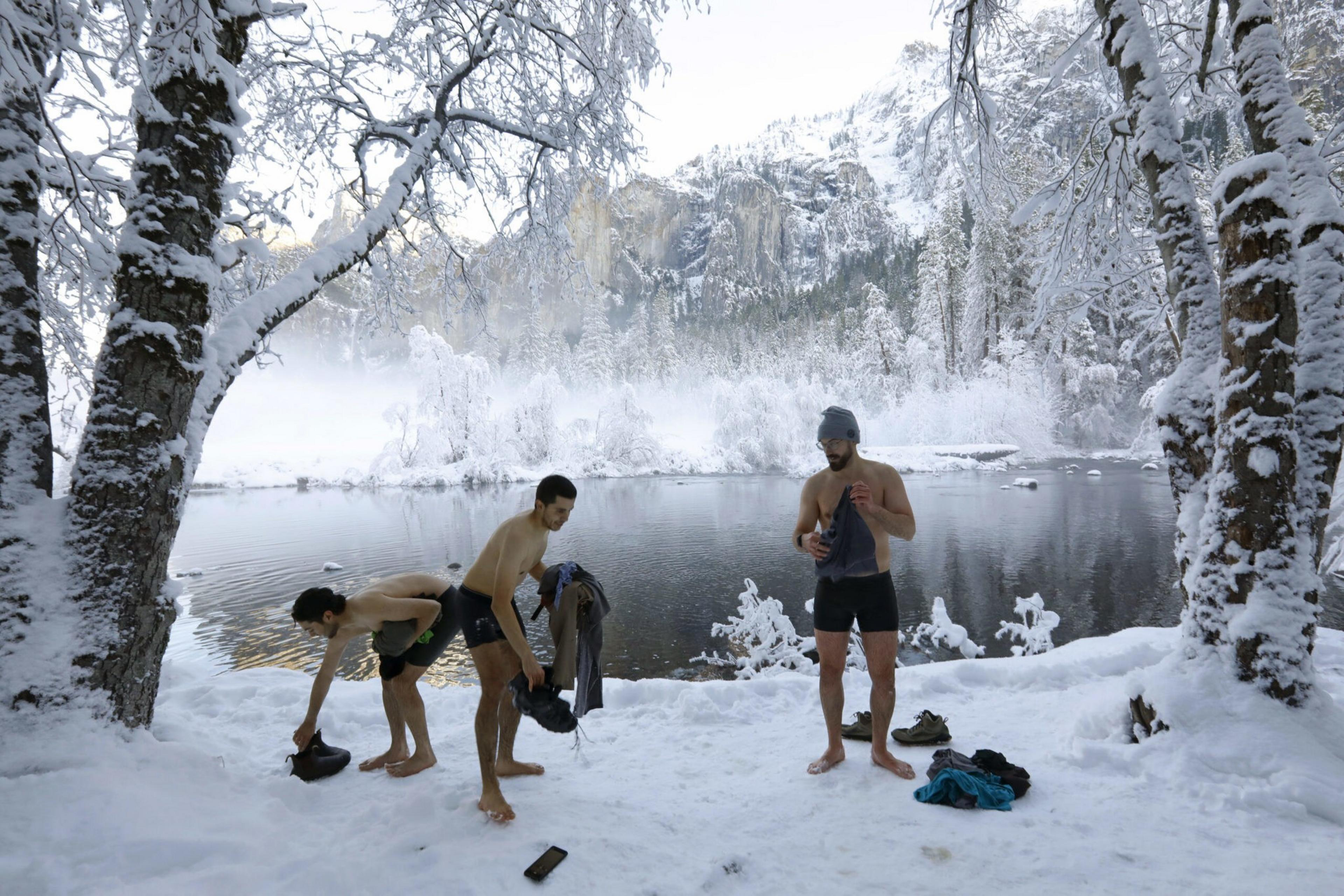 Three shirtless men in shorts stand barefoot on snowy ground beside a icy lake surrounded by snow-covered trees and mountains.