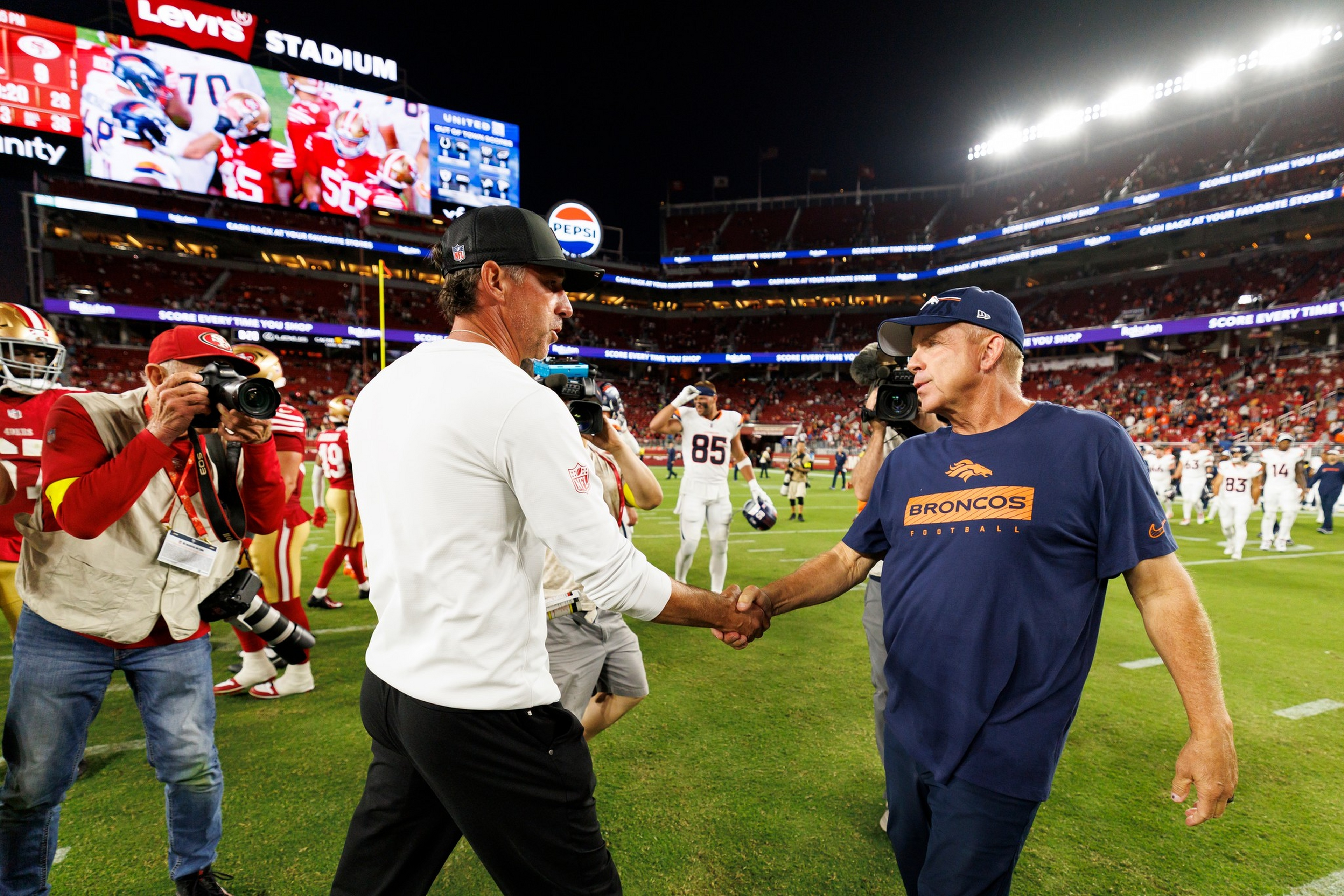 Two football coaches shake hands on the field at Levi's Stadium after a game, while photographers and players are visible in the background.