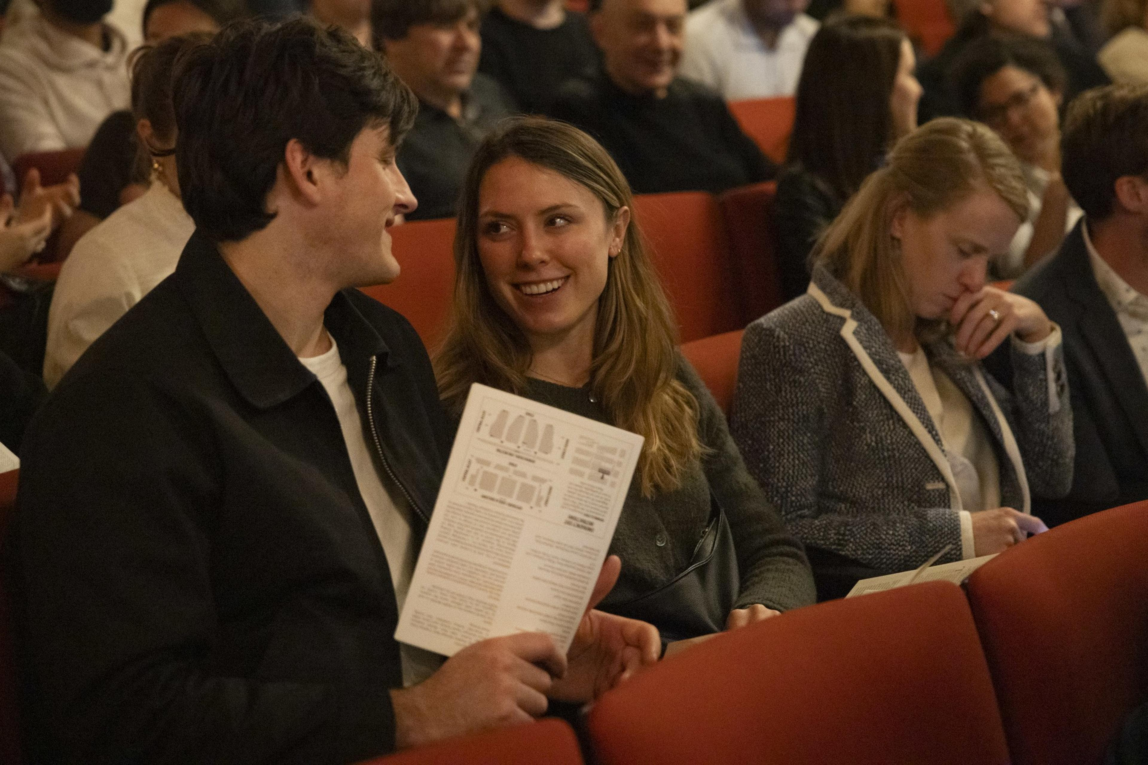 A man and woman sit in a theater, smiling at each other, holding a program. A woman next to them looks down at her own program in a thoughtful manner.