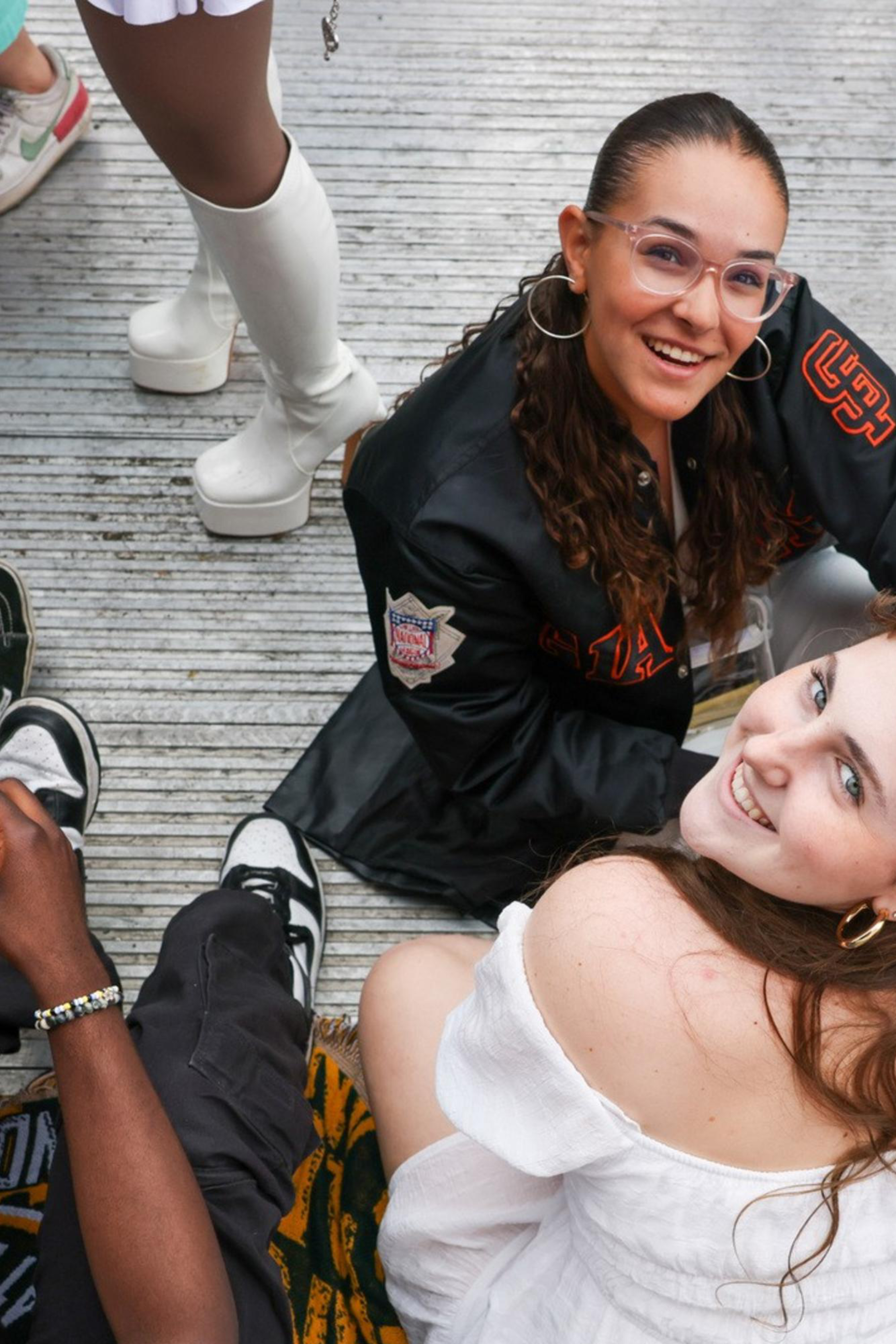 The image shows a group of people sitting on the ground, engaged in various activities. Two women are looking up and smiling towards the camera, while others are using their phones.