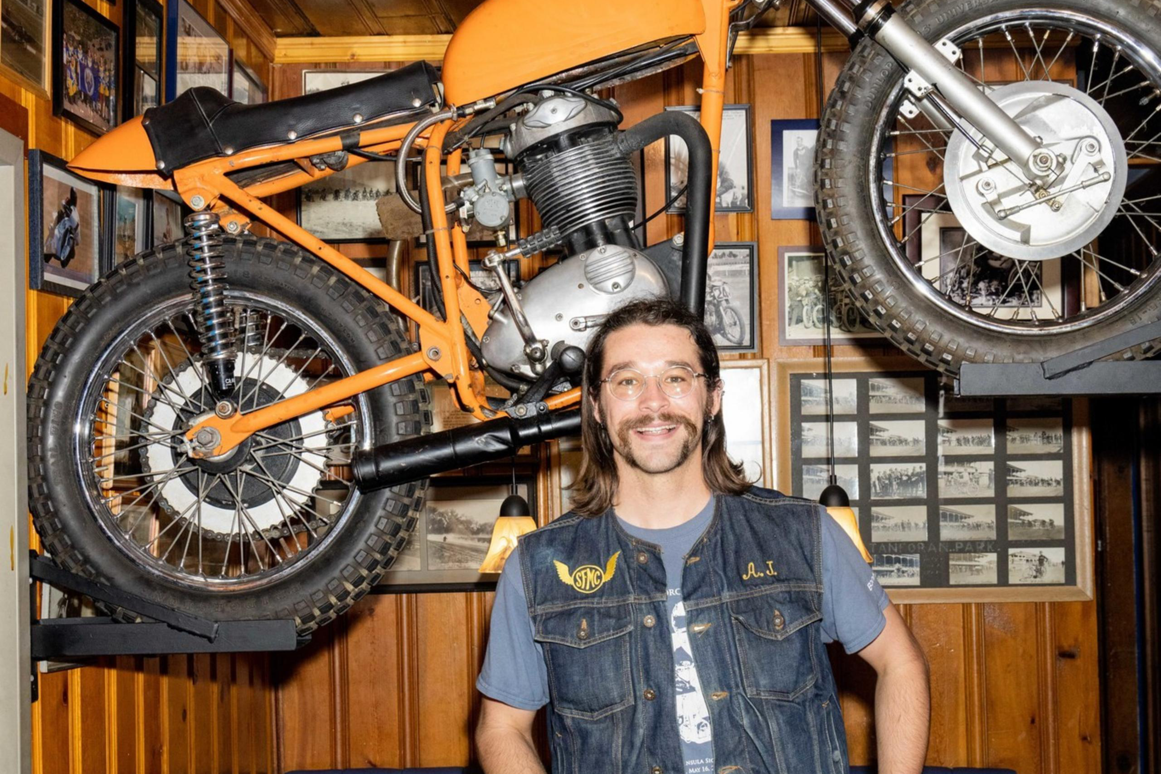 A smiling man with long hair stands holding a bottle, wearing a denim vest and blue jeans. Behind him, an orange motorcycle is mounted on a wall with pictures.