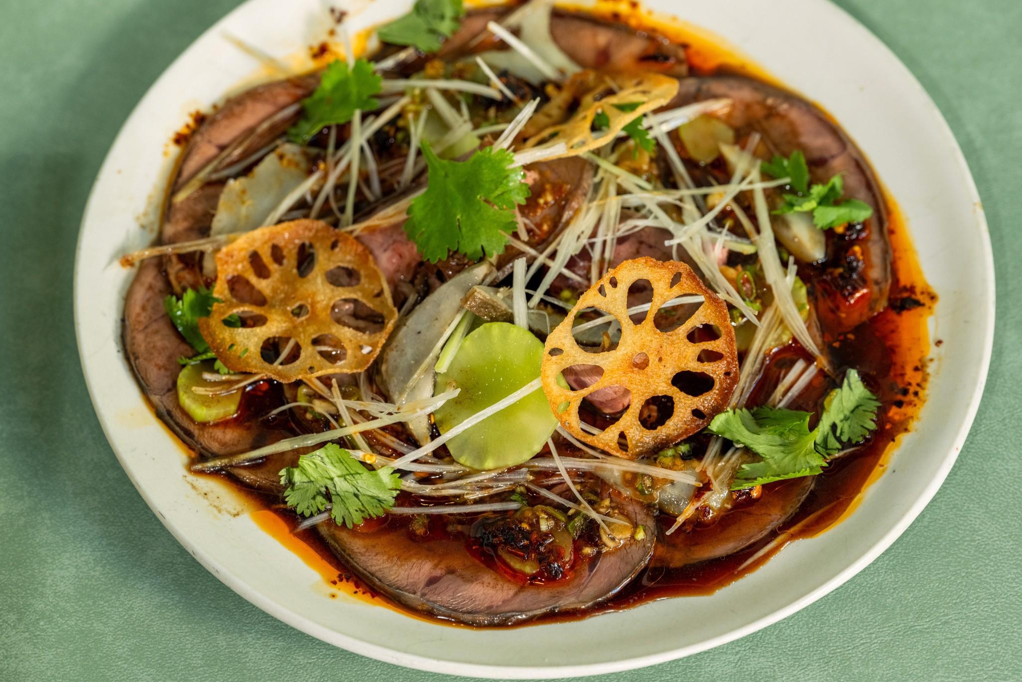 Sliced beef in chili oil topped with lotus root chips, cilantro, thinly sliced ginger, and green vegetables on a white plate.