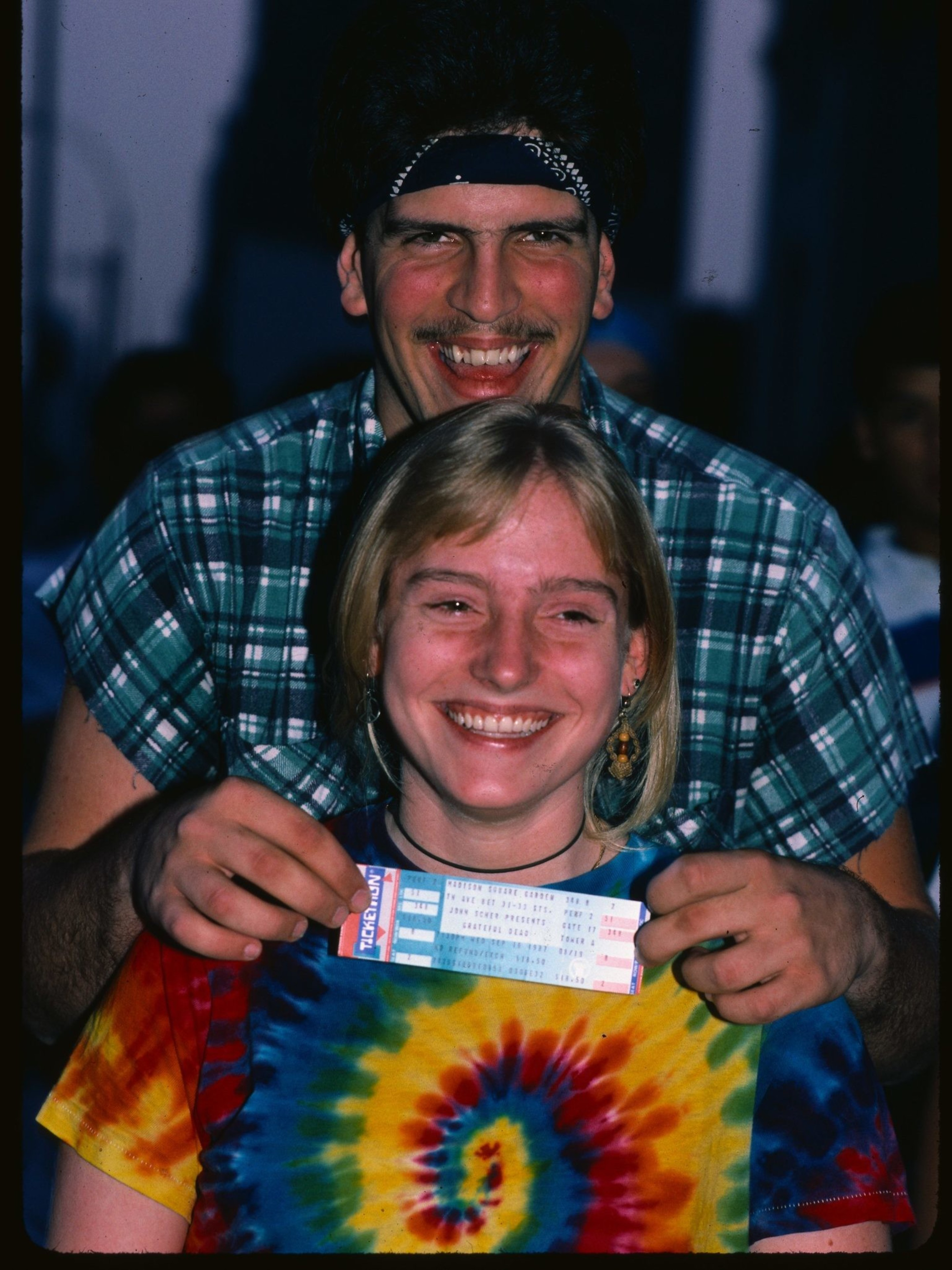 A smiling woman in a tie-dye shirt holds tickets in front of her. A man in a plaid shirt with a bandana leans over her, grinning widely.