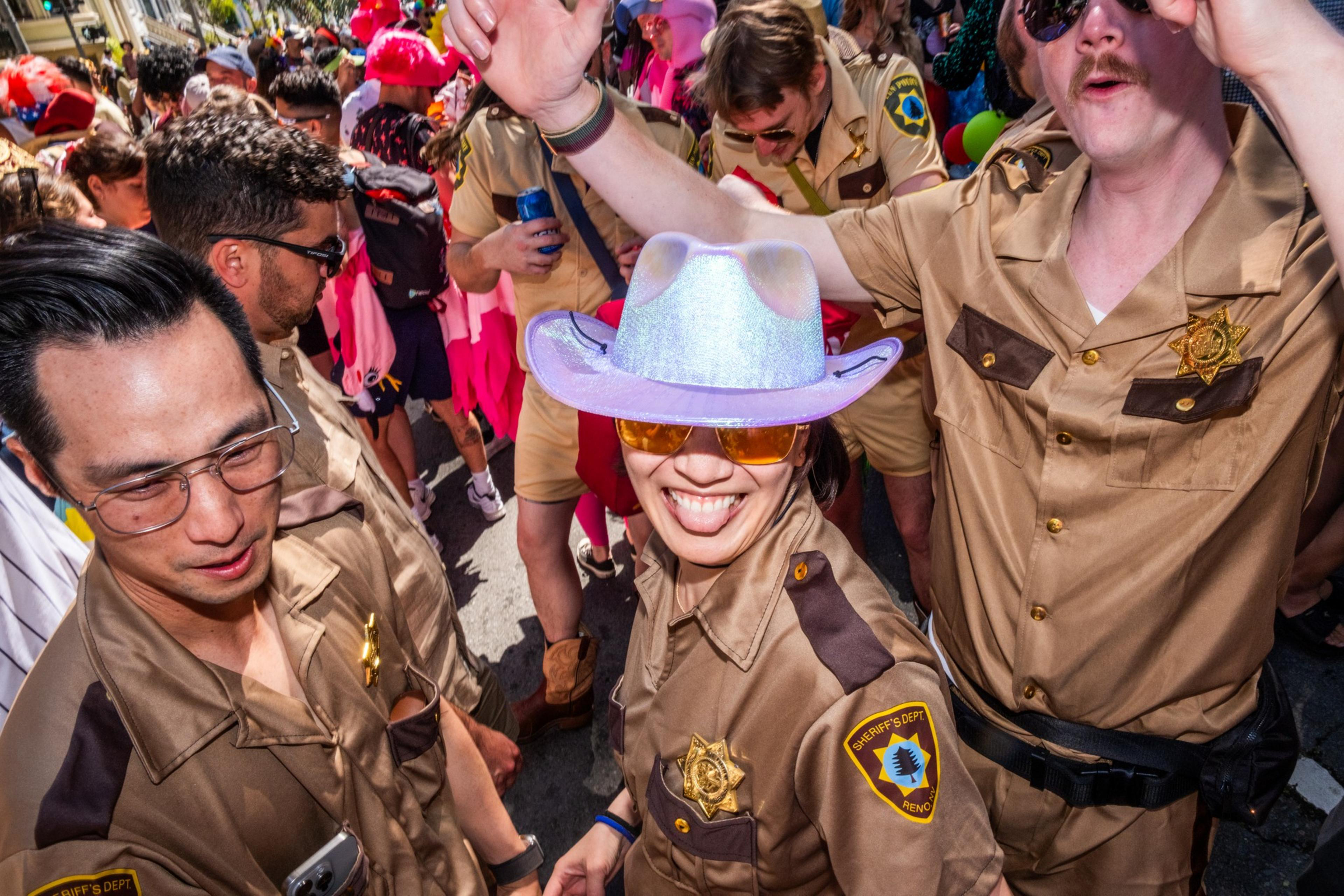 A group of people is dressed in tan sheriff uniforms with badges, smiling and interacting at a lively outdoor event. One wears a shiny cowboy hat.