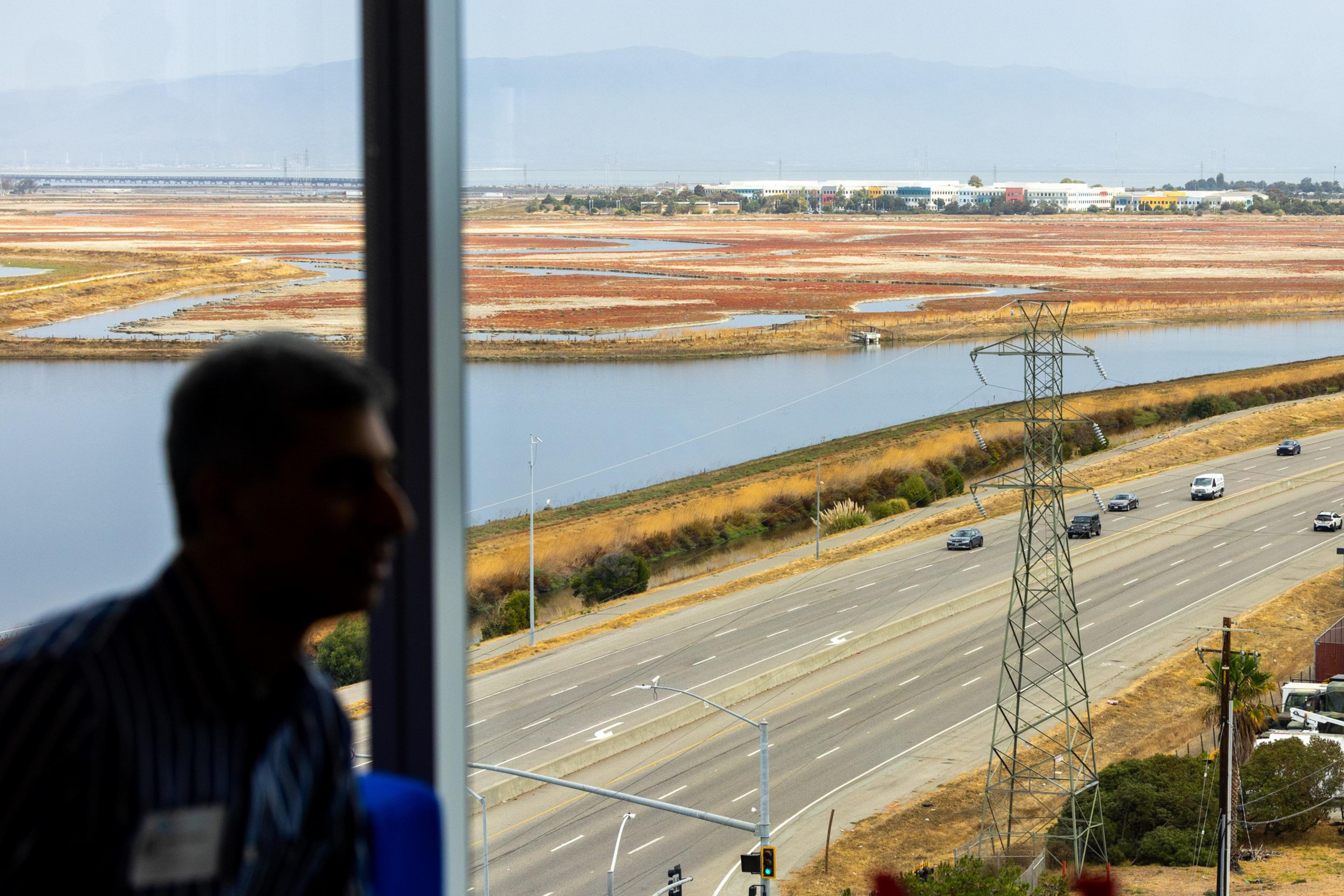A man’s silhouette is seen inside, looking out a window at a highway with cars, a power line tower, water channels, and red vegetation under a hazy sky.