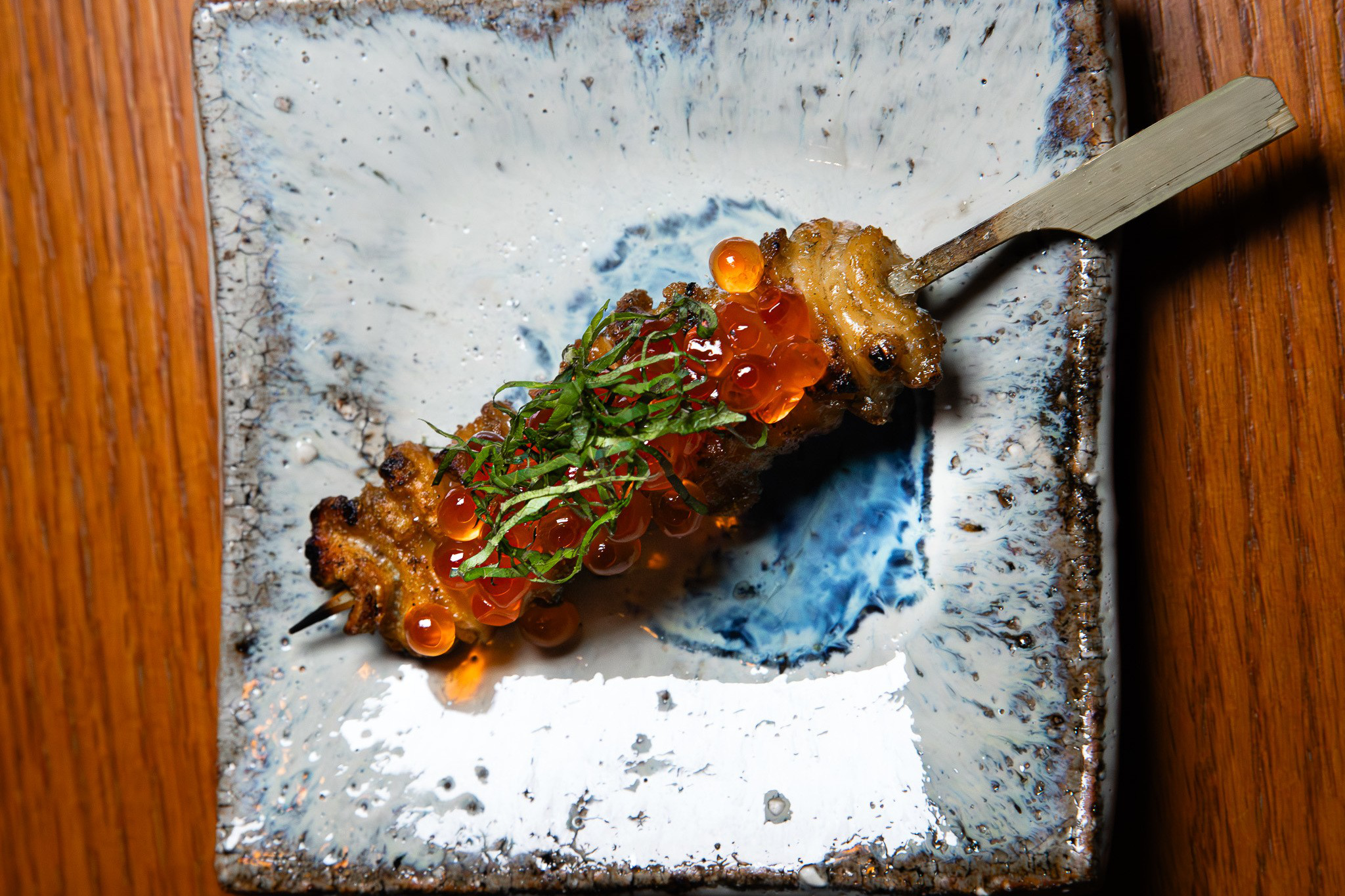 A grilled skewer topped with bright orange salmon roe and thinly sliced green herbs rests on a square ceramic plate with a blue and white glaze.