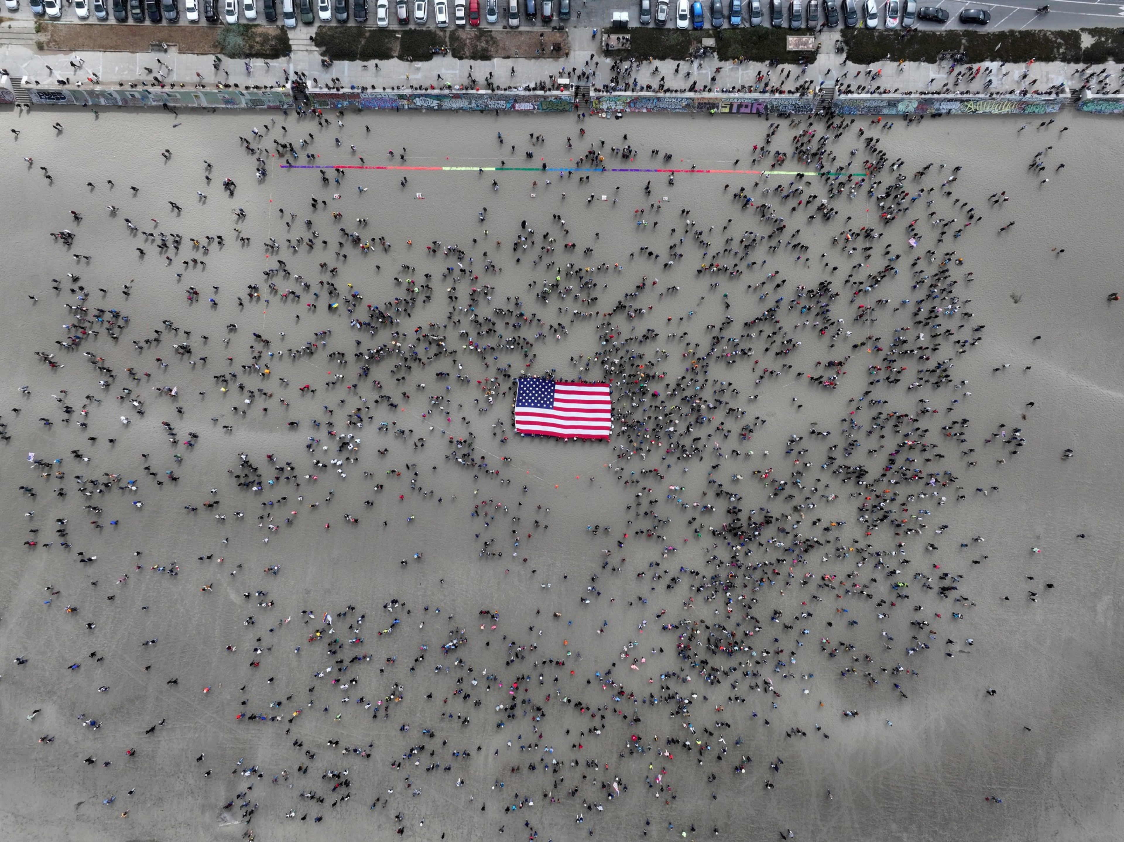 A large crowd of people is gathered on a beach around an American flag. The flag is in the center, and parked cars line the top of the image.