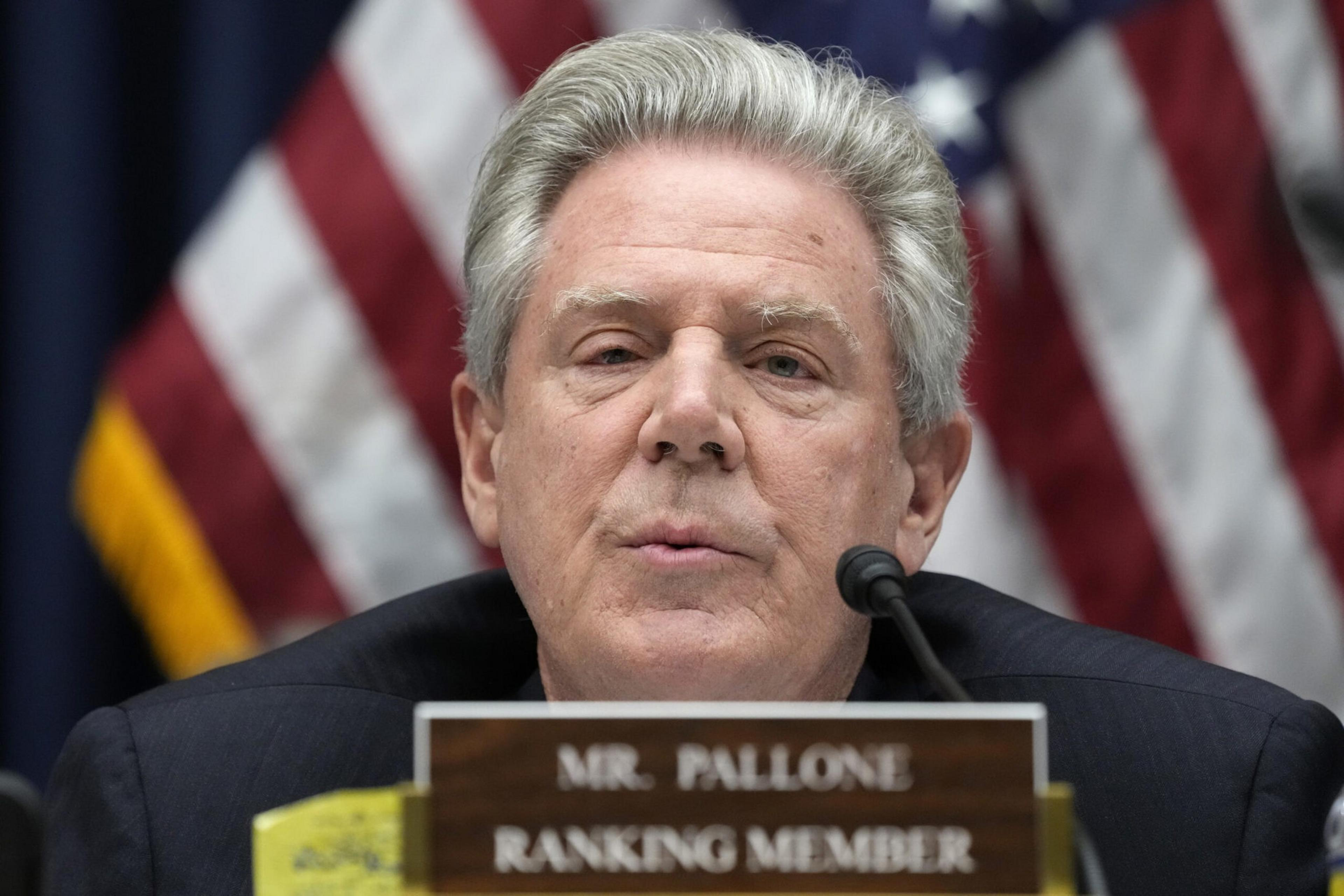 A man with gray hair speaks into a microphone behind a nameplate that reads "Mr. Pallone Ranking Member," with American flags in the background.