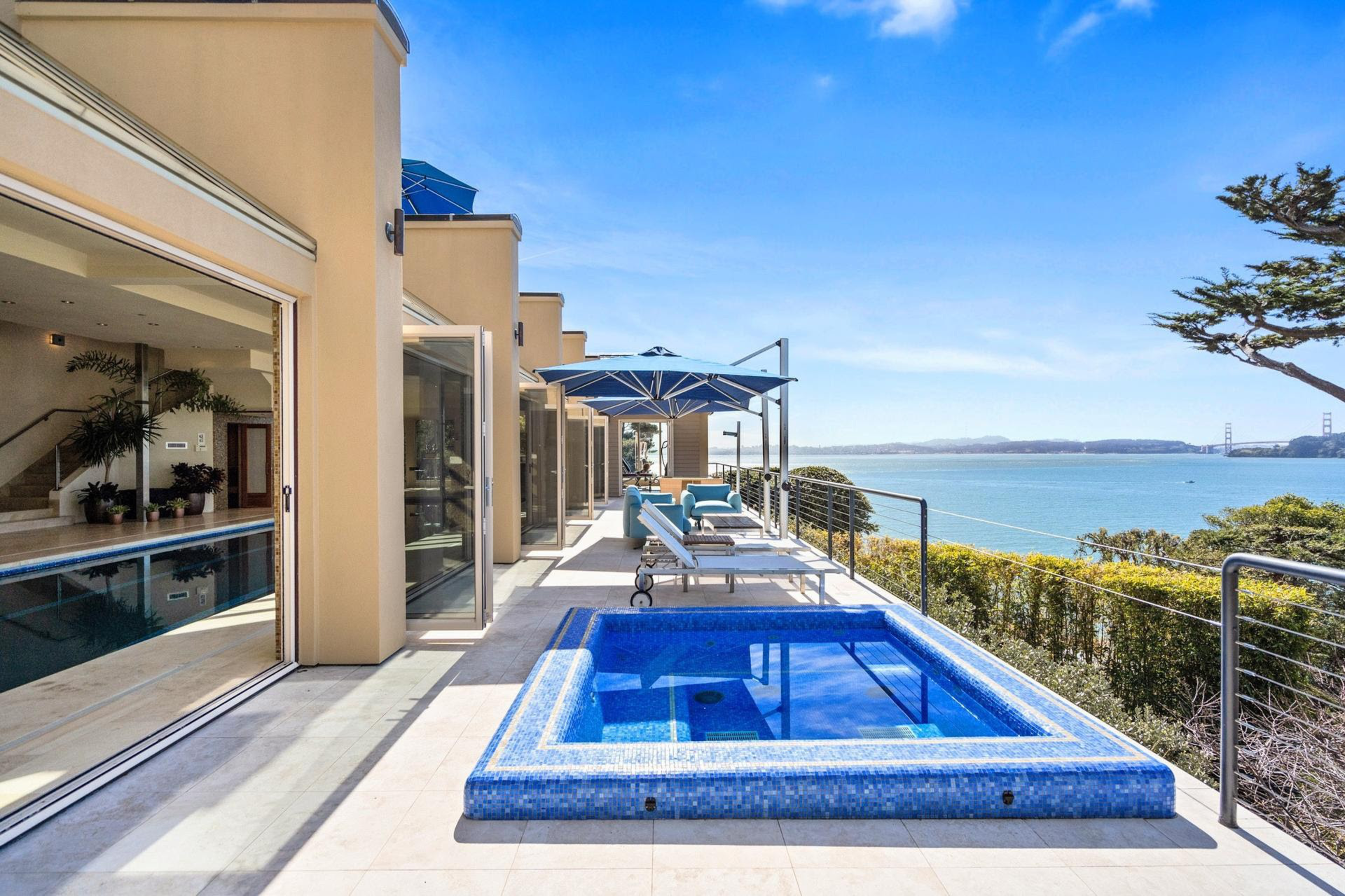 A modern outdoor patio features a square blue-tiled hot tub, lounge chairs with blue umbrellas, and a view of water and the Golden Gate Bridge.