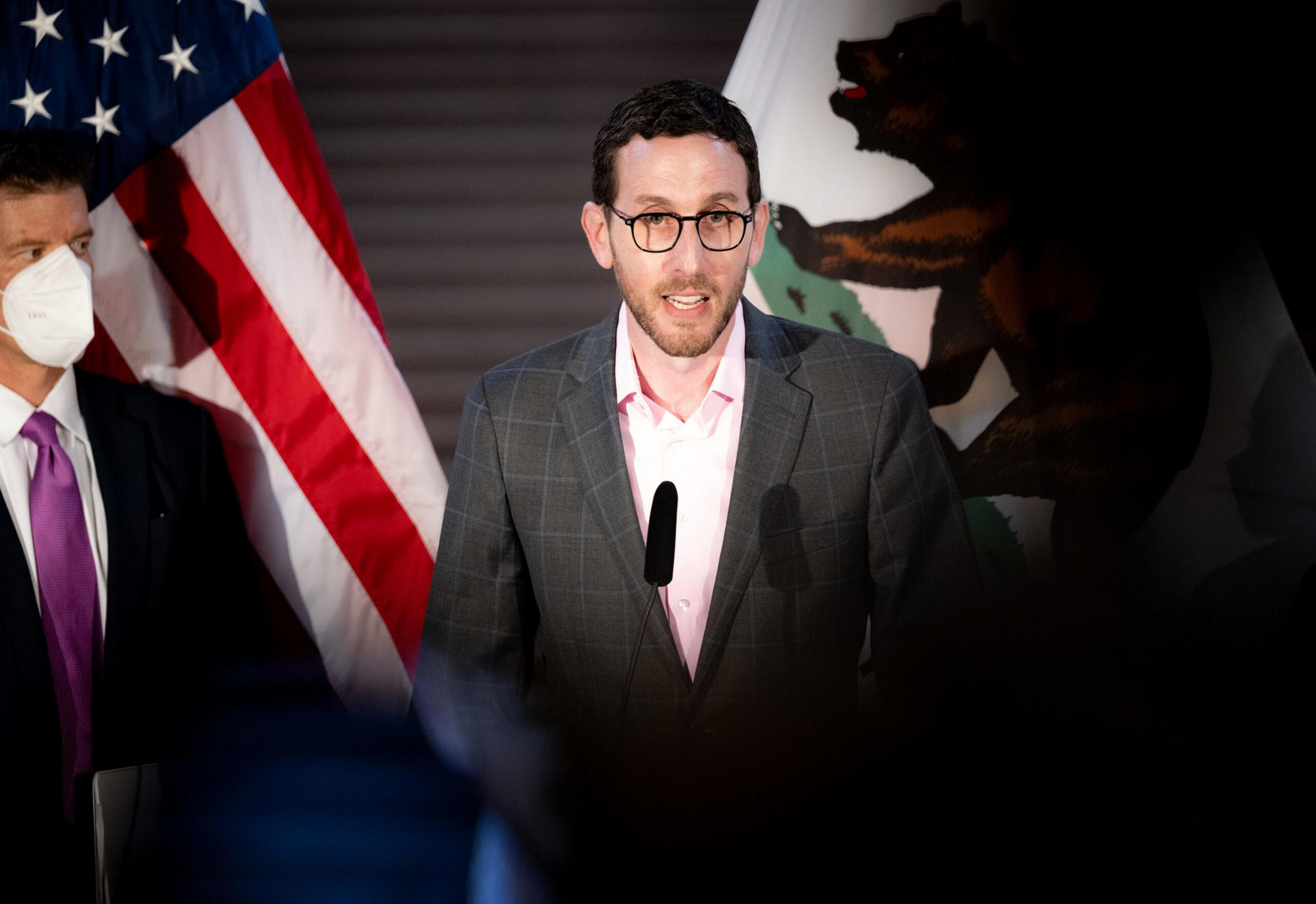 A man wearing glasses and a gray plaid blazer speaks at a podium with an American flag and part of a bear flag behind him.