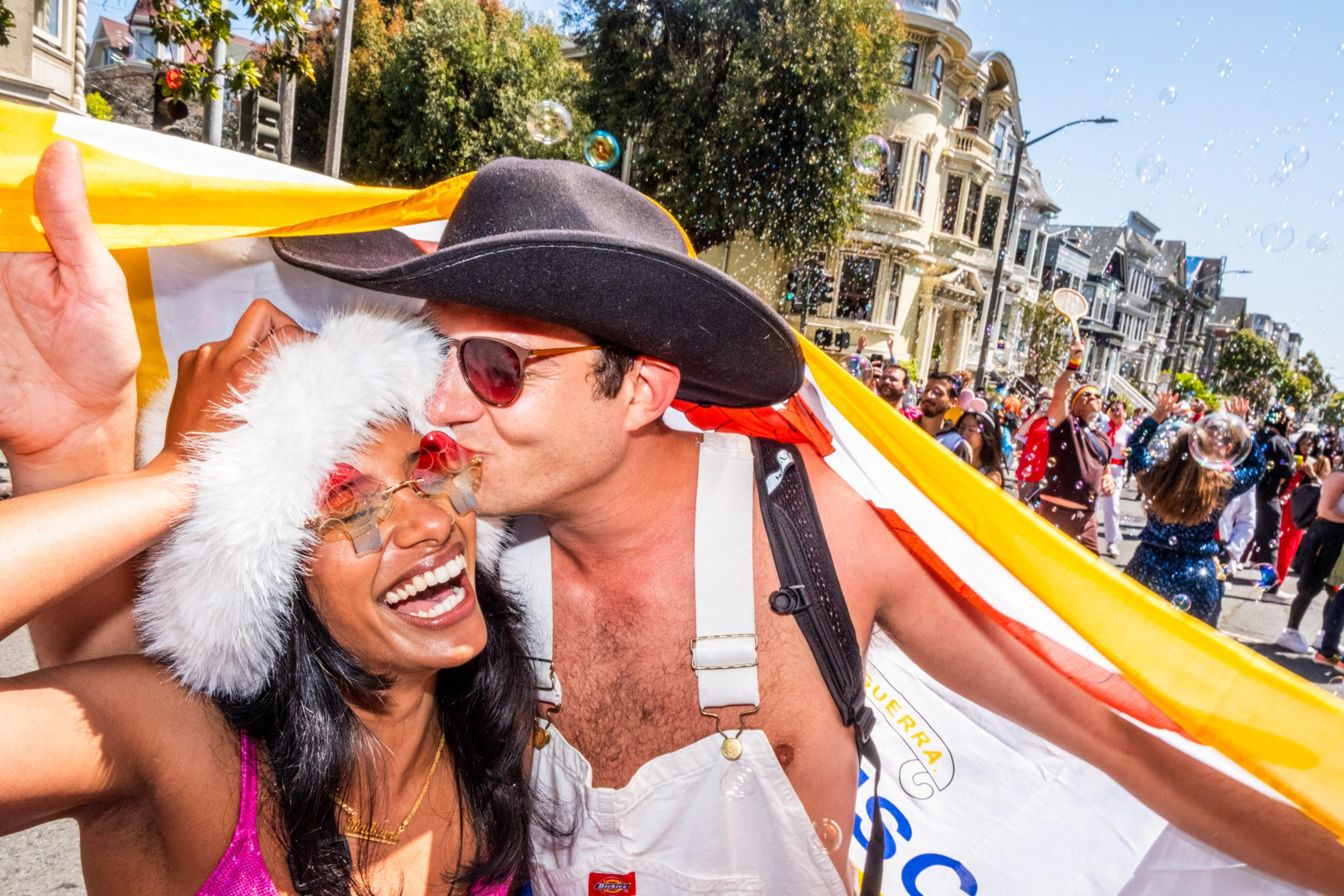 A joyful scene shows two people smiling and sharing a moment in a festive street with colorful outfits and bubbles floating around, surrounded by a lively crowd.