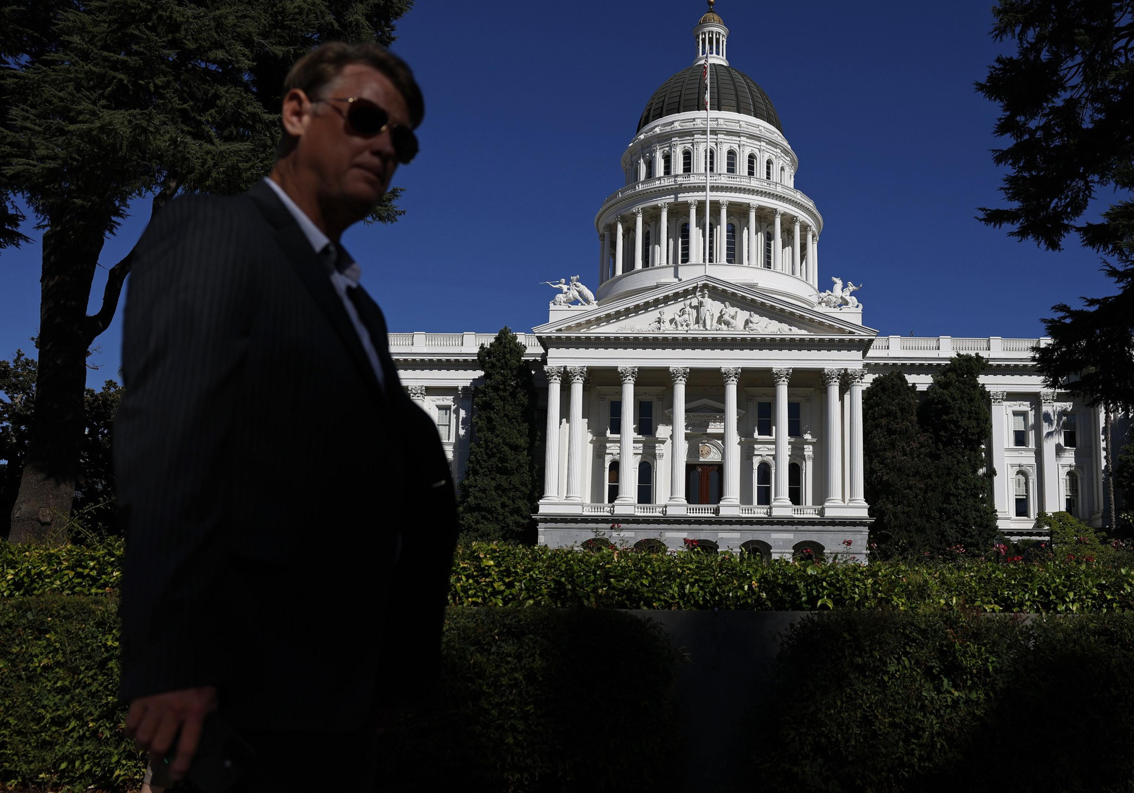 A man wearing sunglasses and a dark suit stands in shadow before the sunlit white dome and columns of a classical government building.