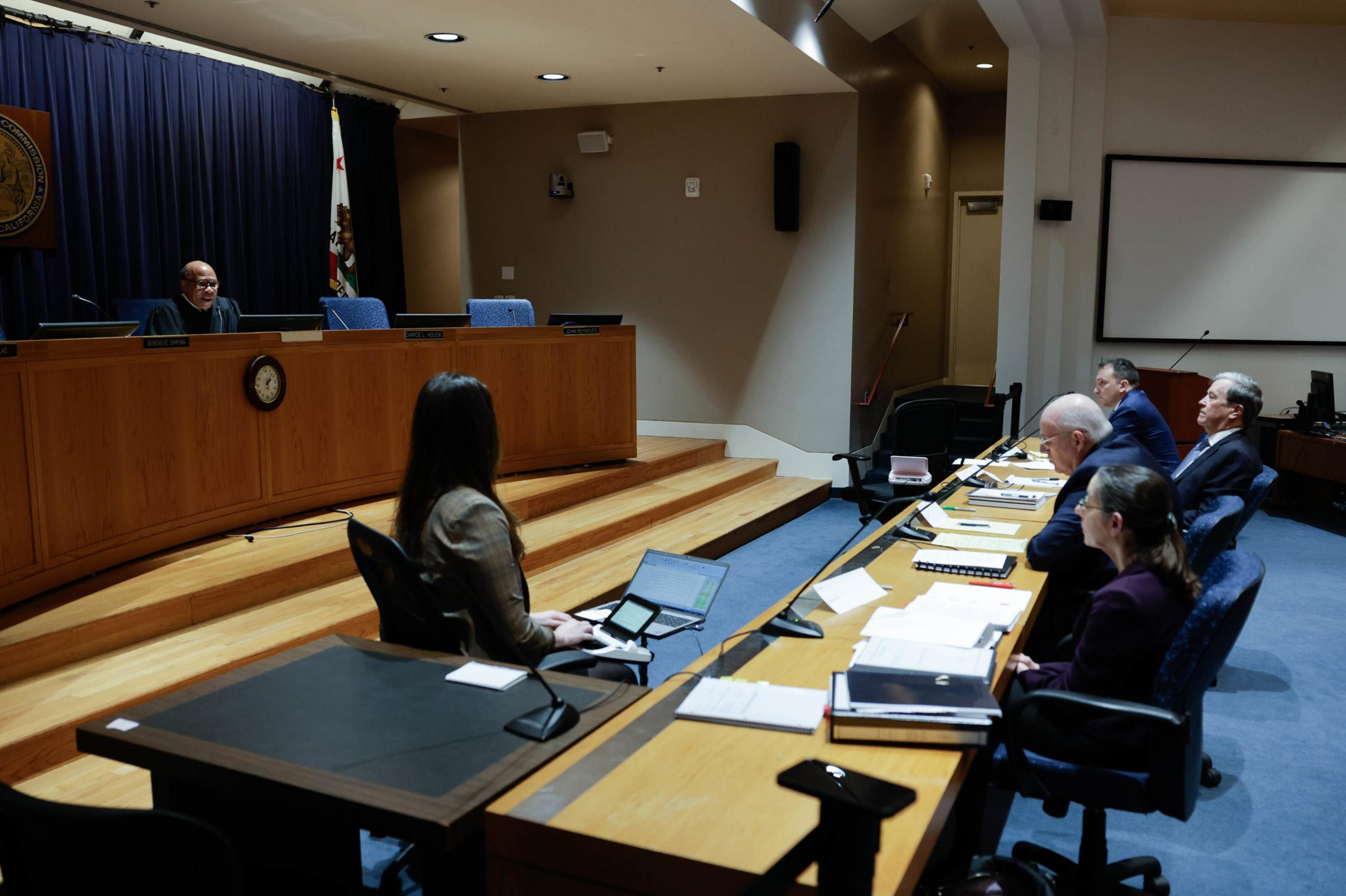 A courtroom scene with a judge presiding and a woman facing a row of seated individuals, possibly lawyers.