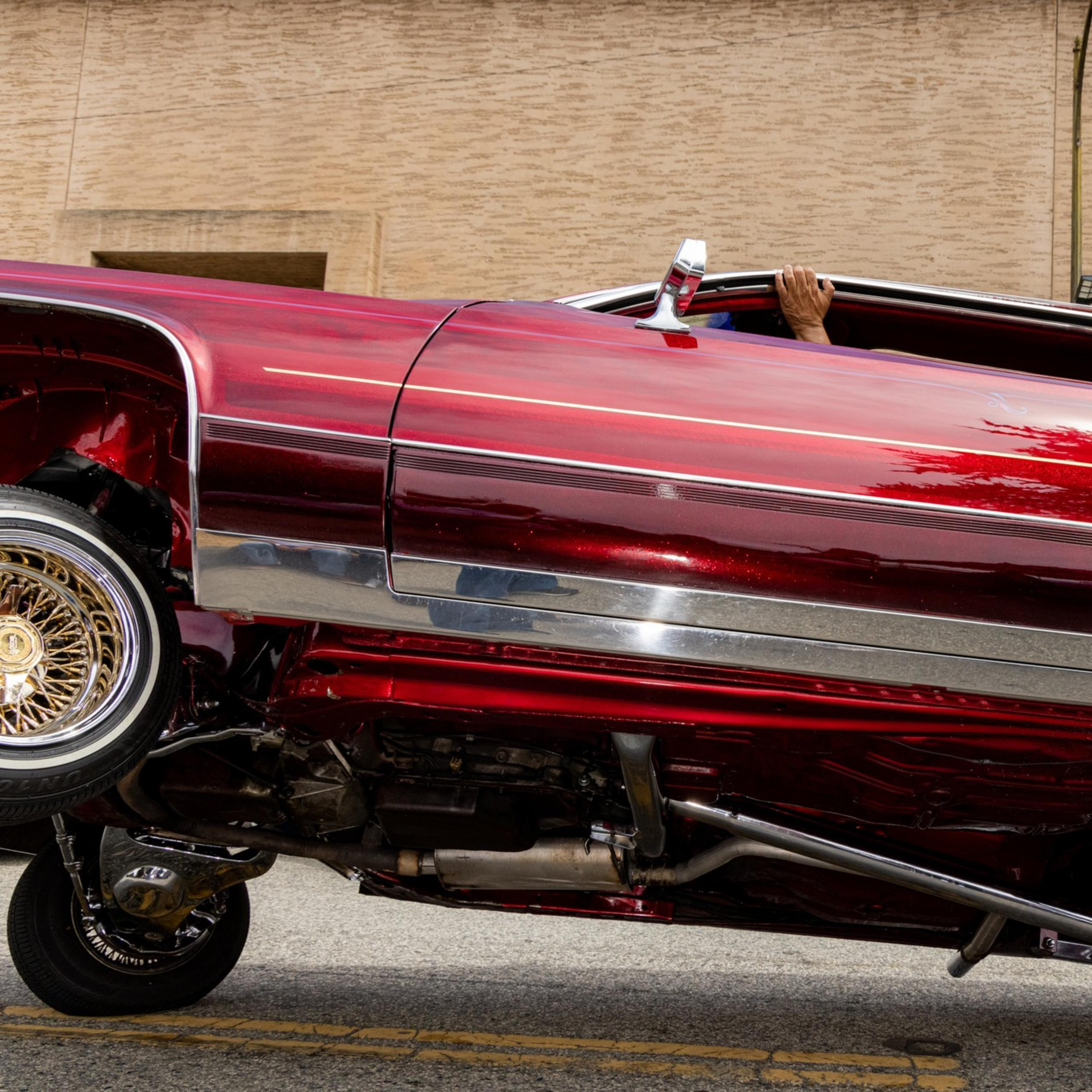 A red lowrider car with shiny chrome details is tilted diagonally, showing its undercarriage. A hand is visible from the rolled-down window, gripping it.