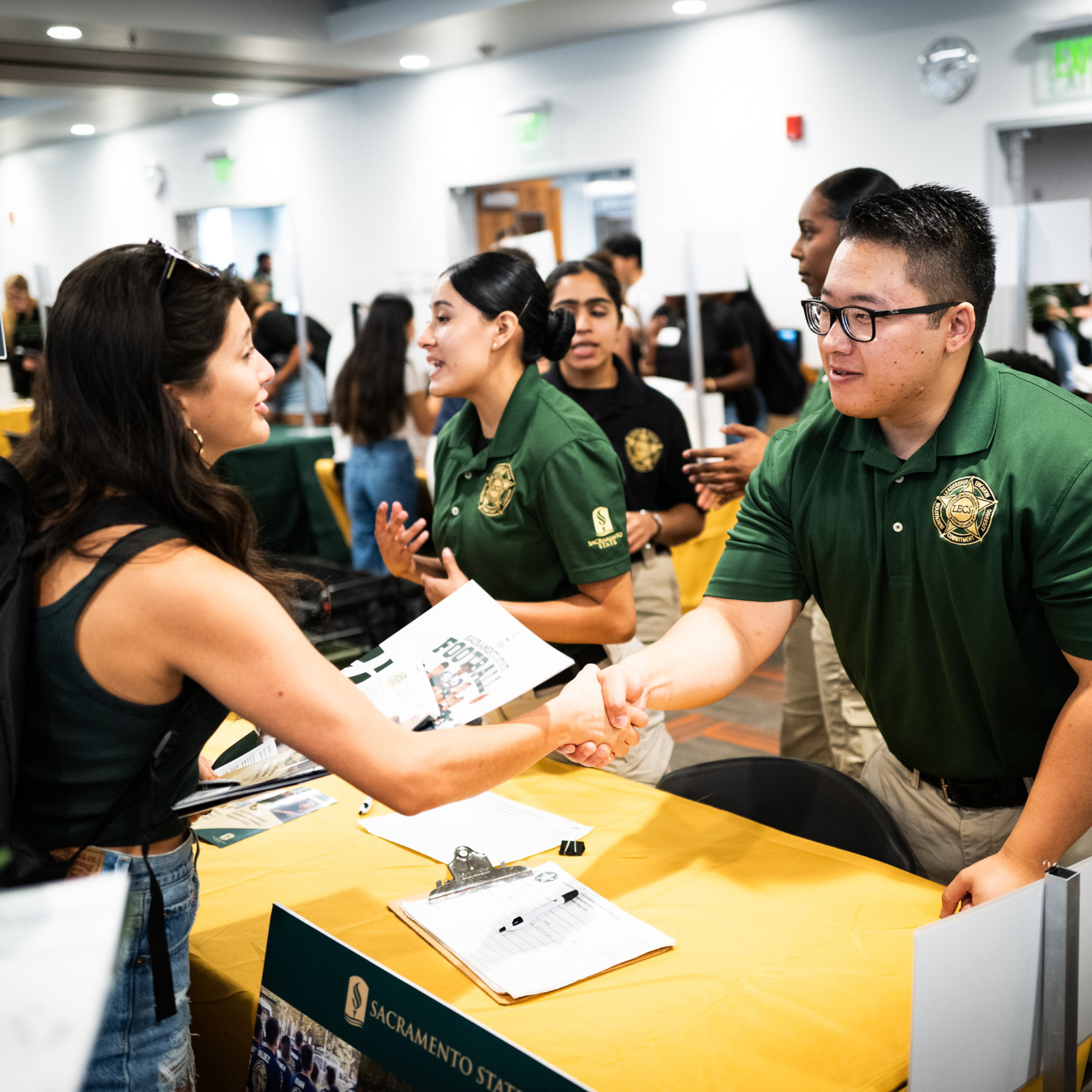 A woman is shaking hands with a man in a green uniform at a booth in a busy indoor event, while others nearby are engaged in conversation.