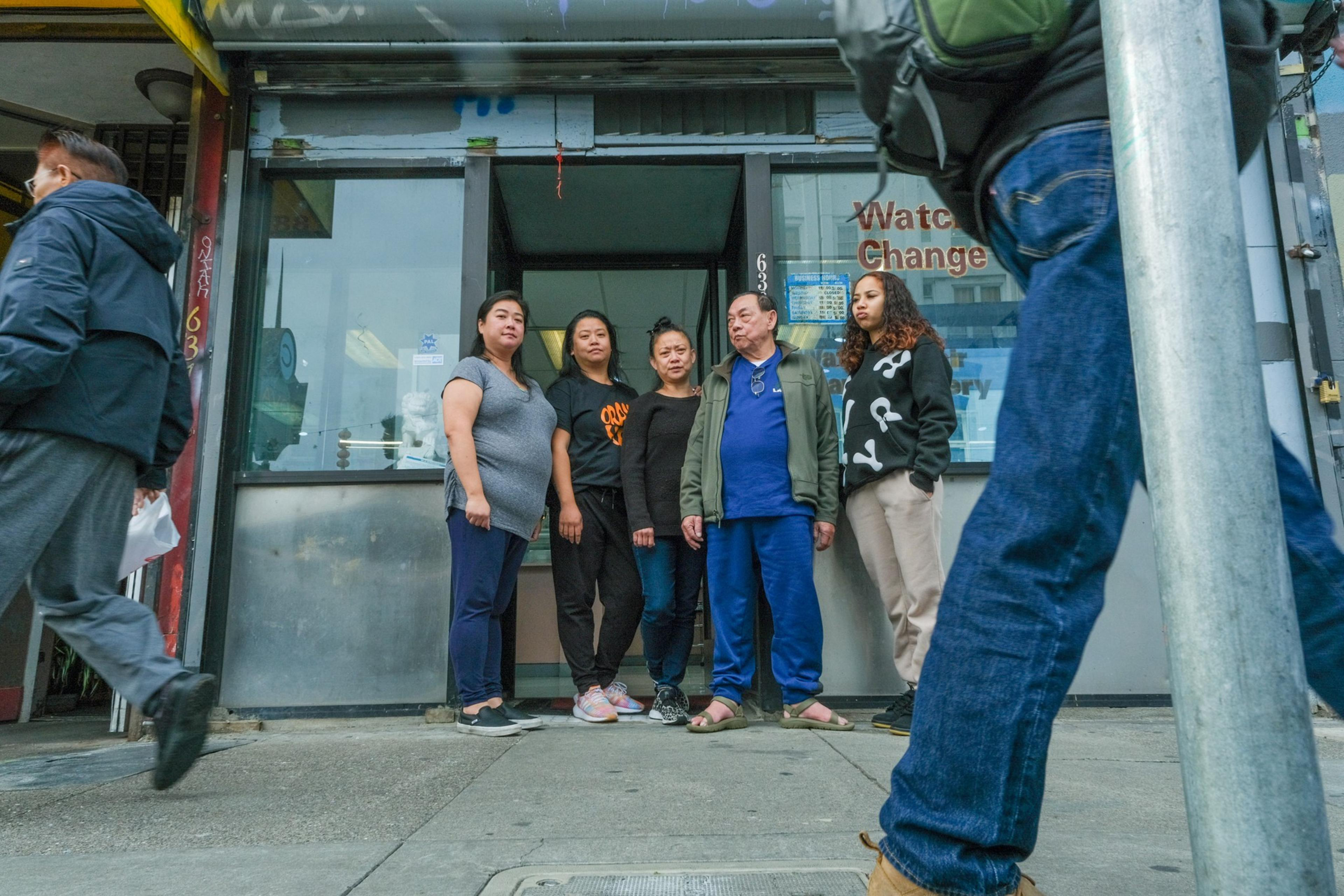 Five adults stand in front of a store, with pedestrians passing by on a city sidewalk.