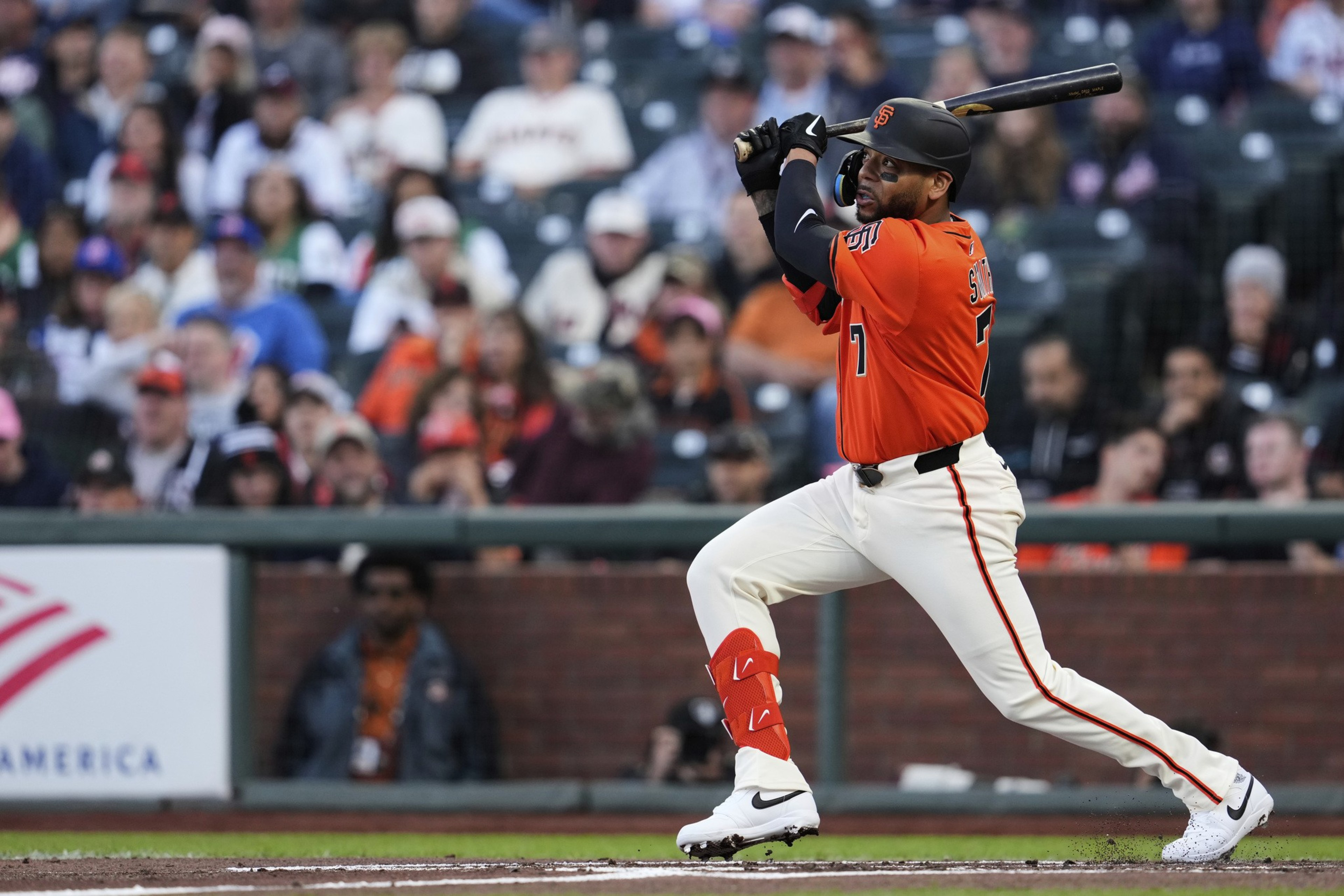 A baseball player in an orange jersey is swinging a bat during a game. The stadium is packed with spectators in the background.
