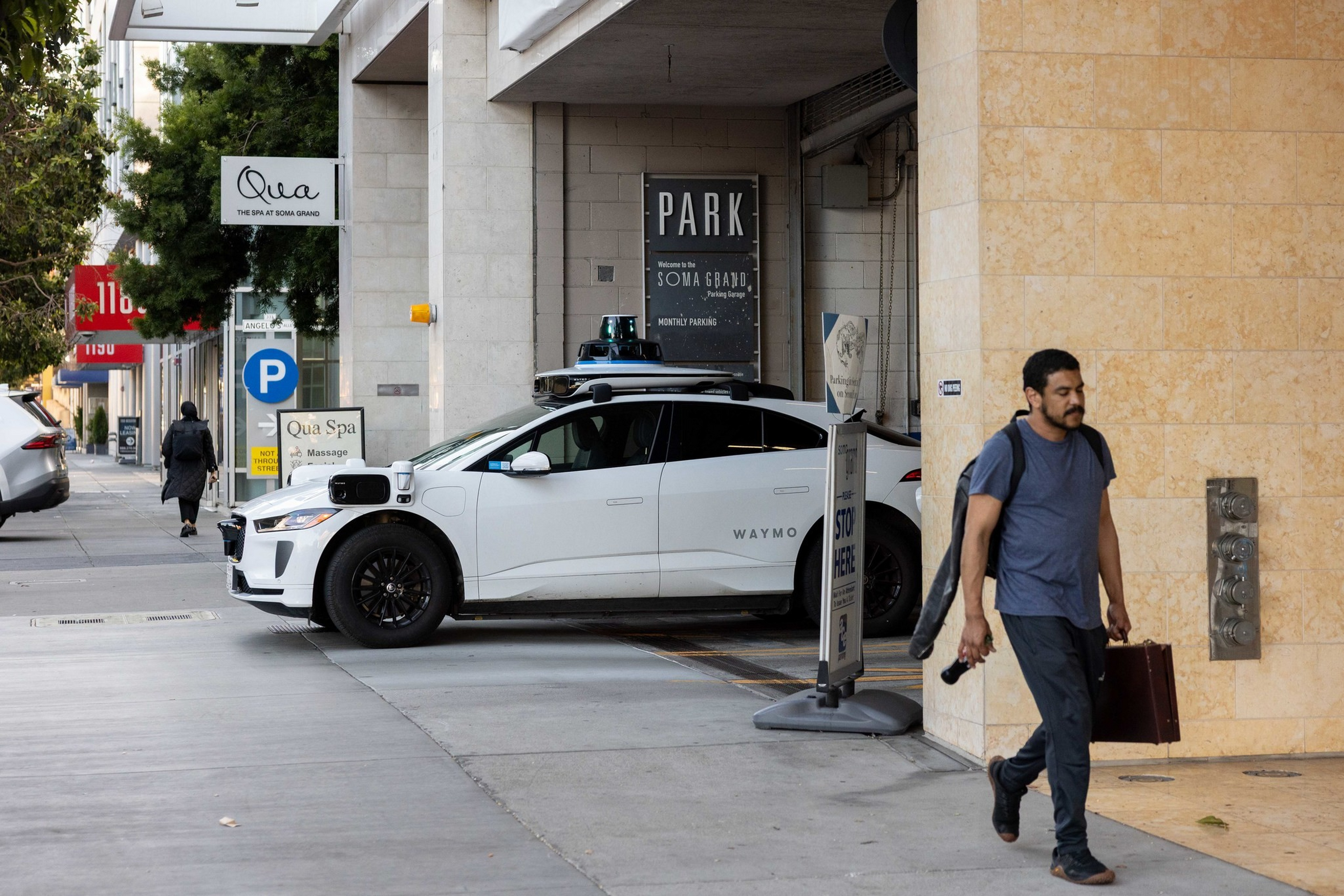 A white Waymo self-driving car is parked near a building entrance, while a man carries a guitar case and briefcase walking nearby.