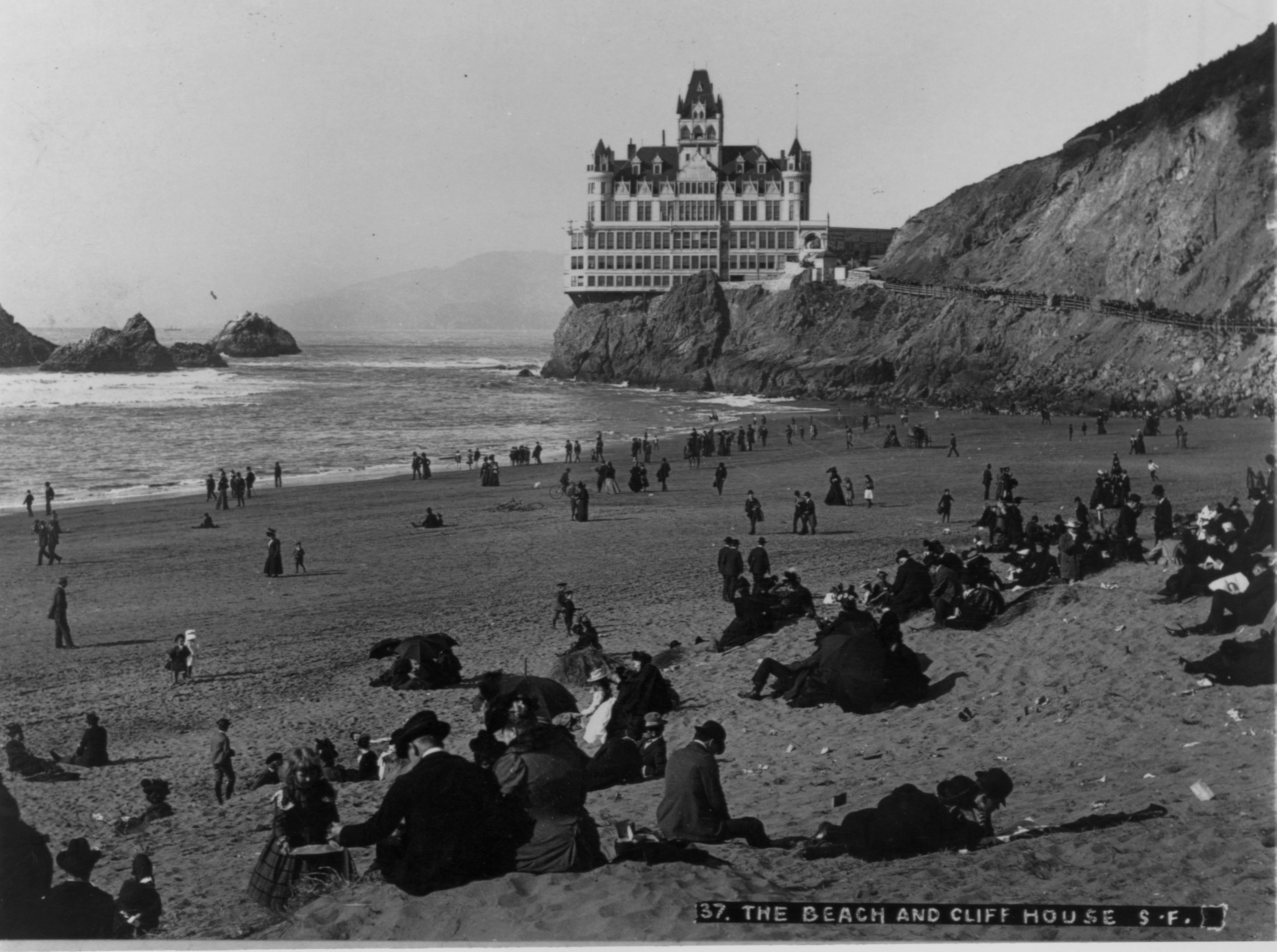 A crowded beach with people dressed in early 1900s attire sits near a large castle-like building atop rocky cliffs by the ocean.