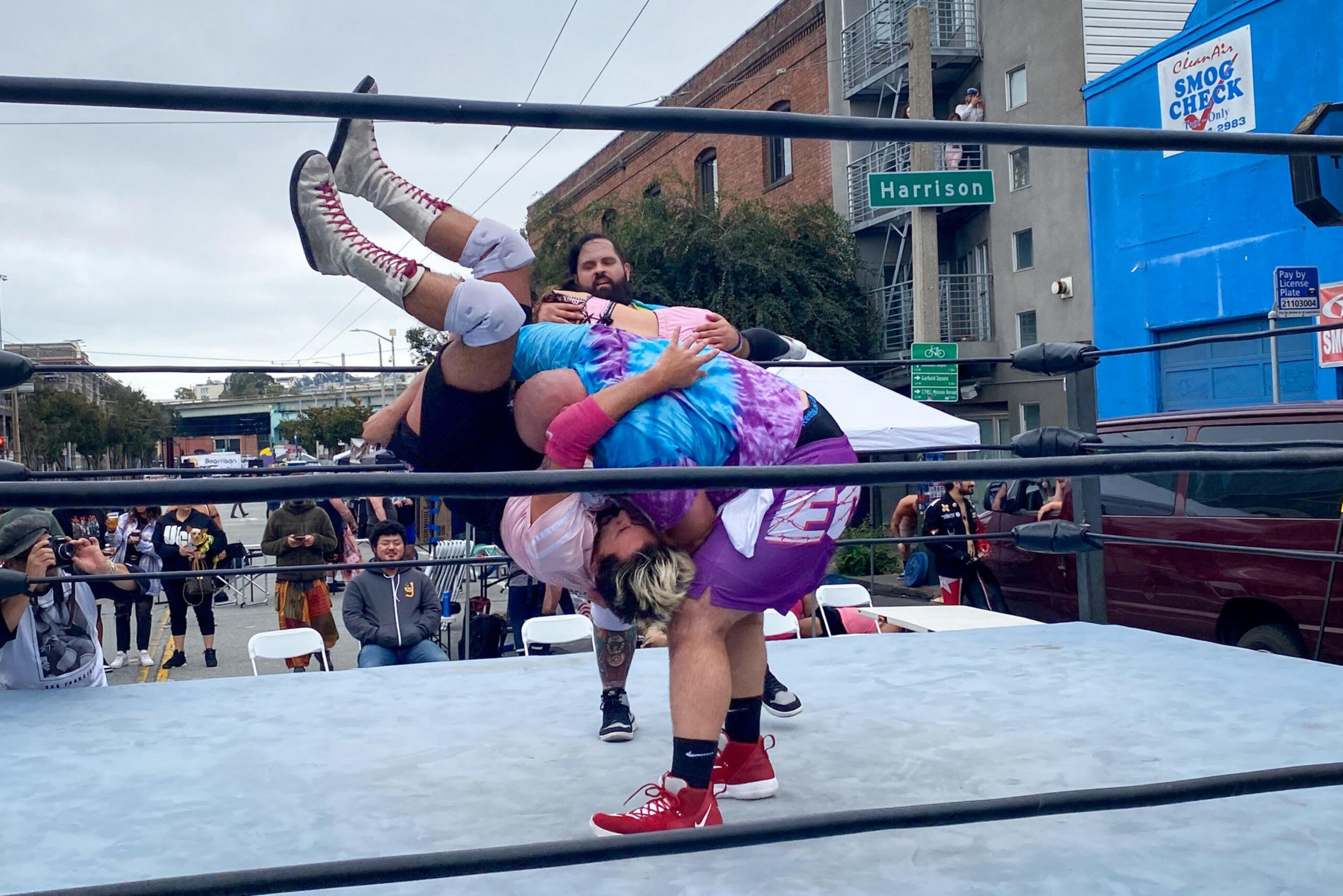A wrestler in purple lifts an opponent upside down in a move inside a street wrestling ring, with a referee and crowd watching nearby.