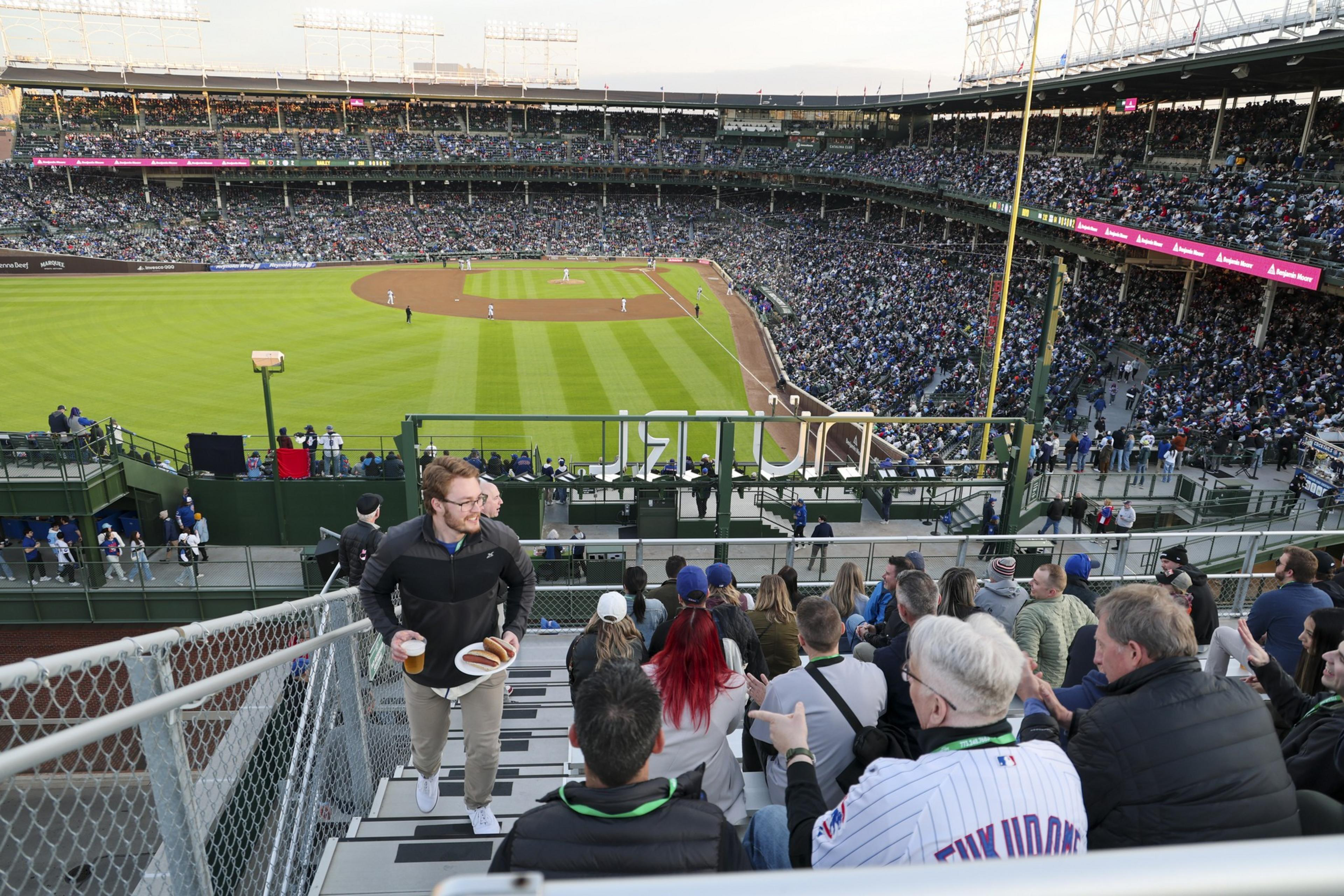 A crowded baseball stadium with a game in progress. Spectators fill the stands, while a man walks upstairs holding food and drinks. The field is brightly lit.