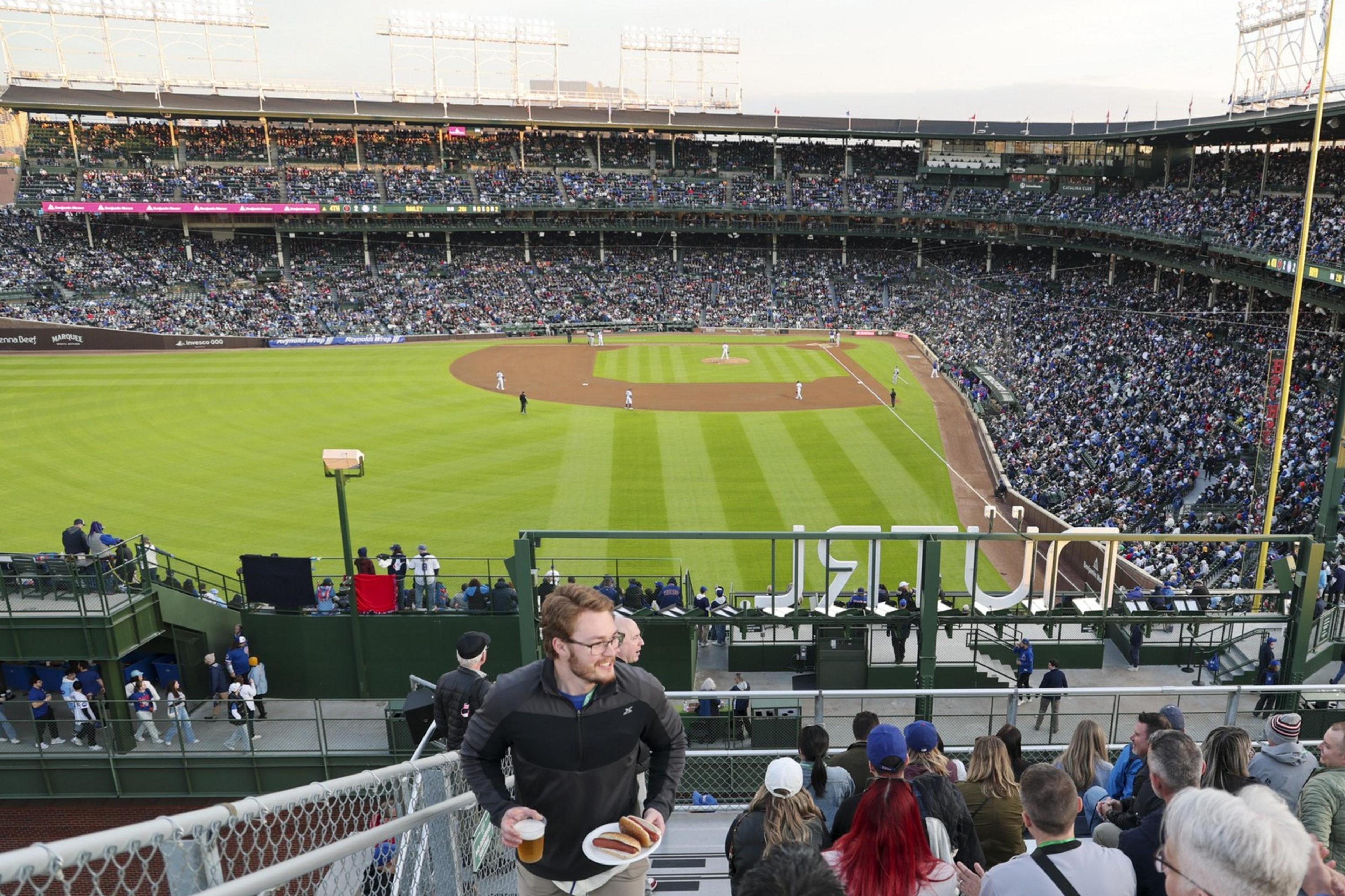 A crowded baseball stadium with a game in progress. Spectators fill the stands, while a man walks upstairs holding food and drinks. The field is brightly lit.