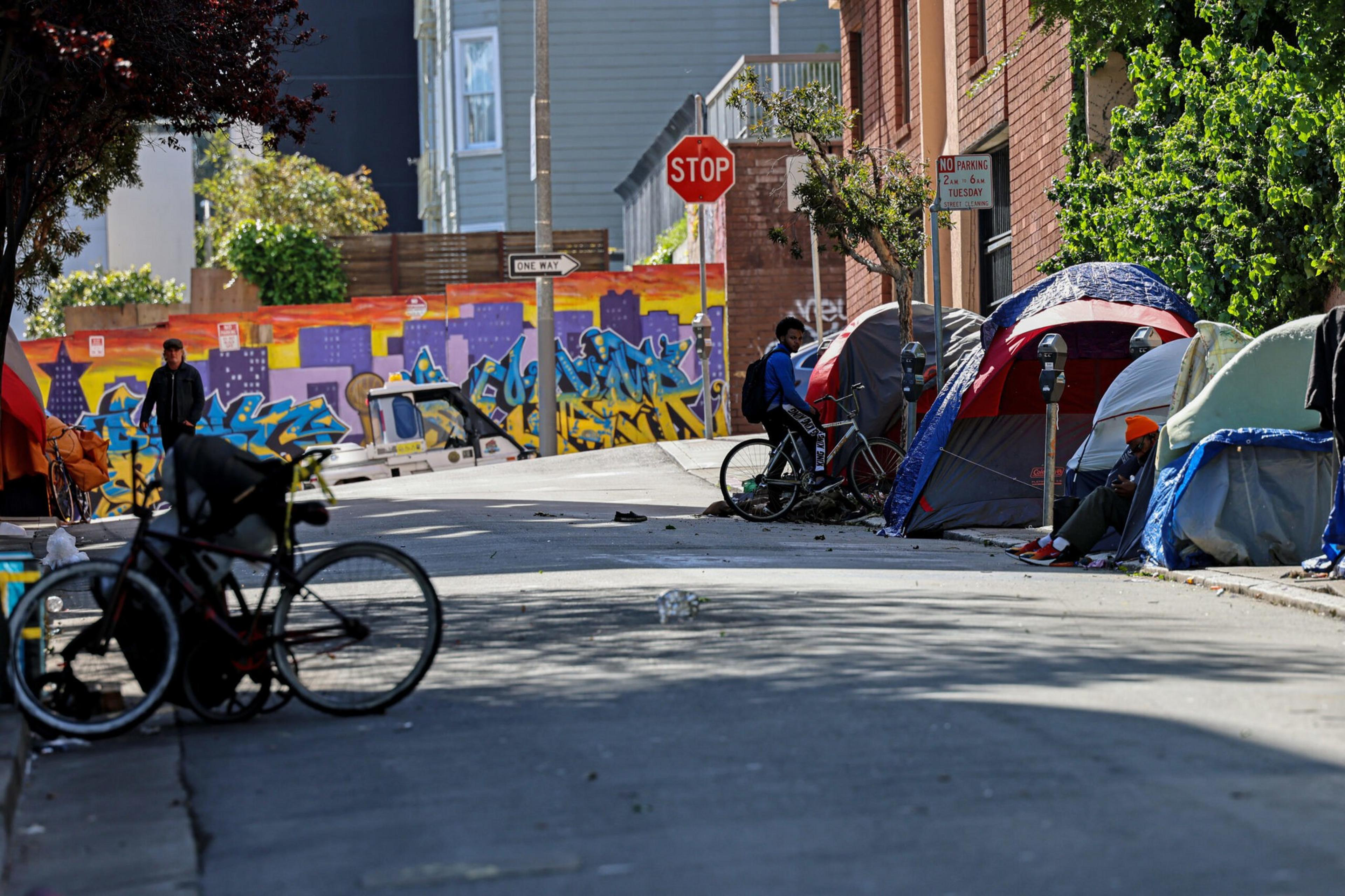 Several tents line a sidewalk beside a street with a stop sign and colorful graffiti on a wall in the background, while people and bicycles occupy the area.