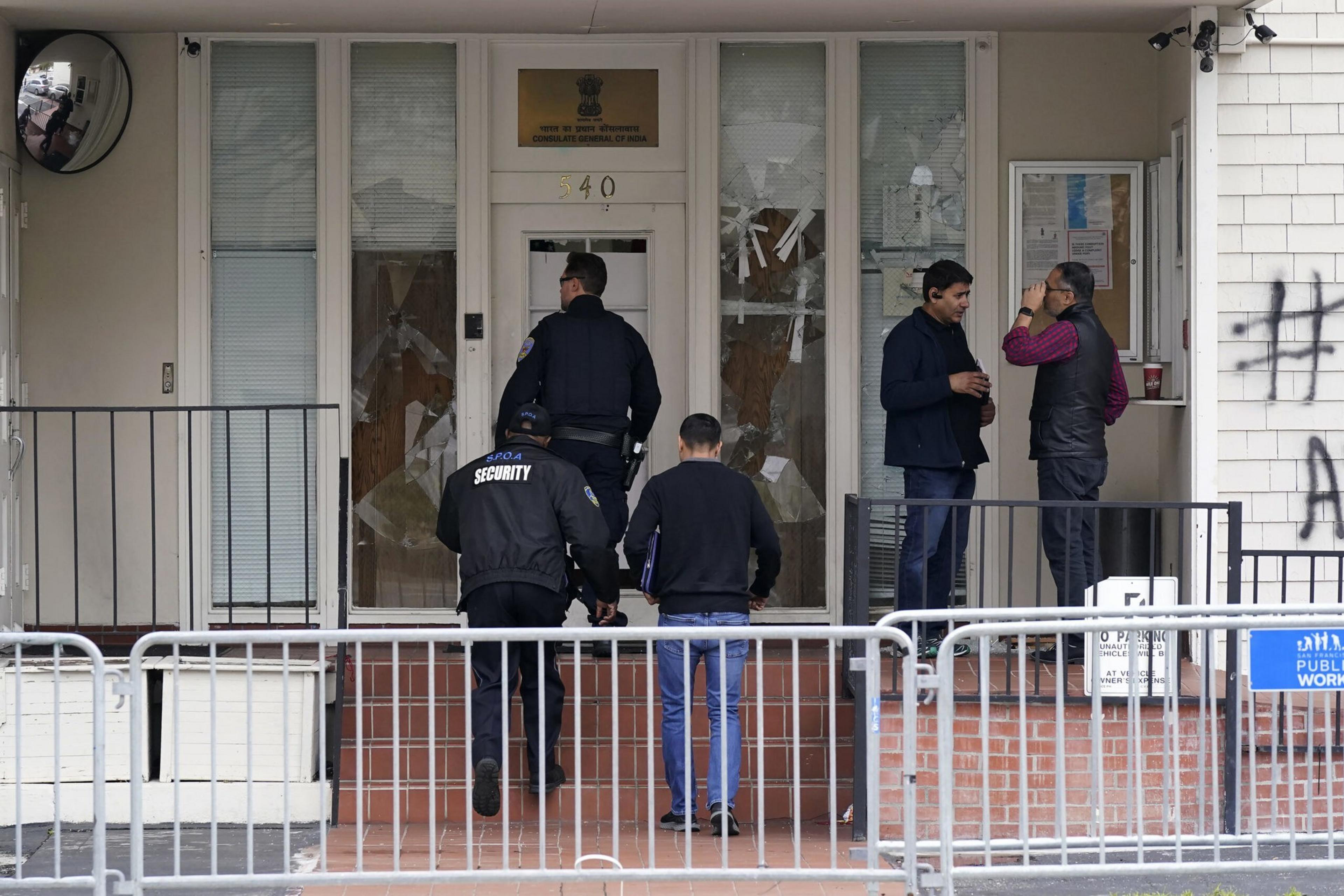 Two men, a security guard, and a police officer enter a building with a sign in Hindi and English, while two others talk near a small window to the right.