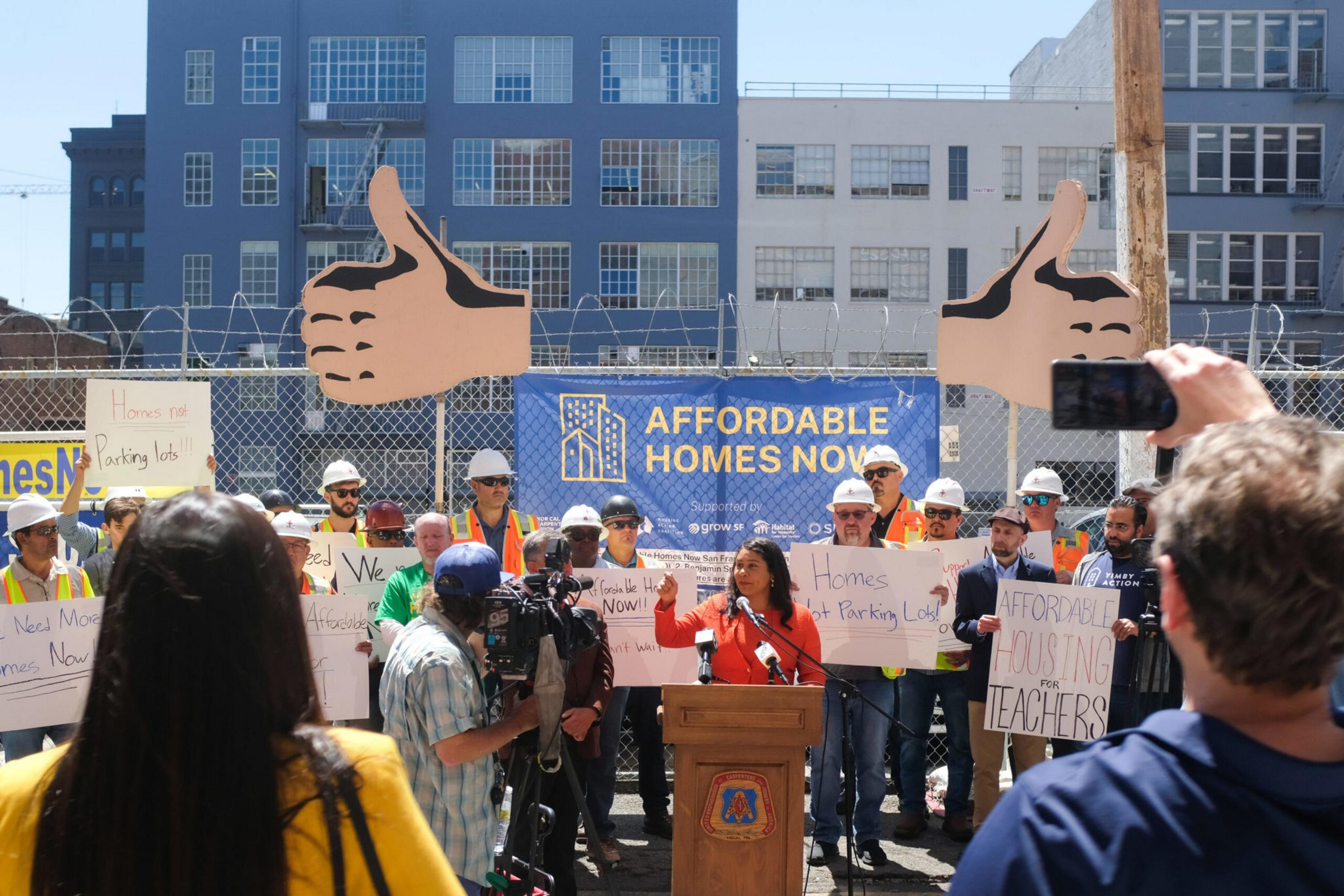 A crowd with signs and helmets holds a rally for affordable homes, with two large thumbs-up signs and a banner behind them reading "Affordable Homes Now."