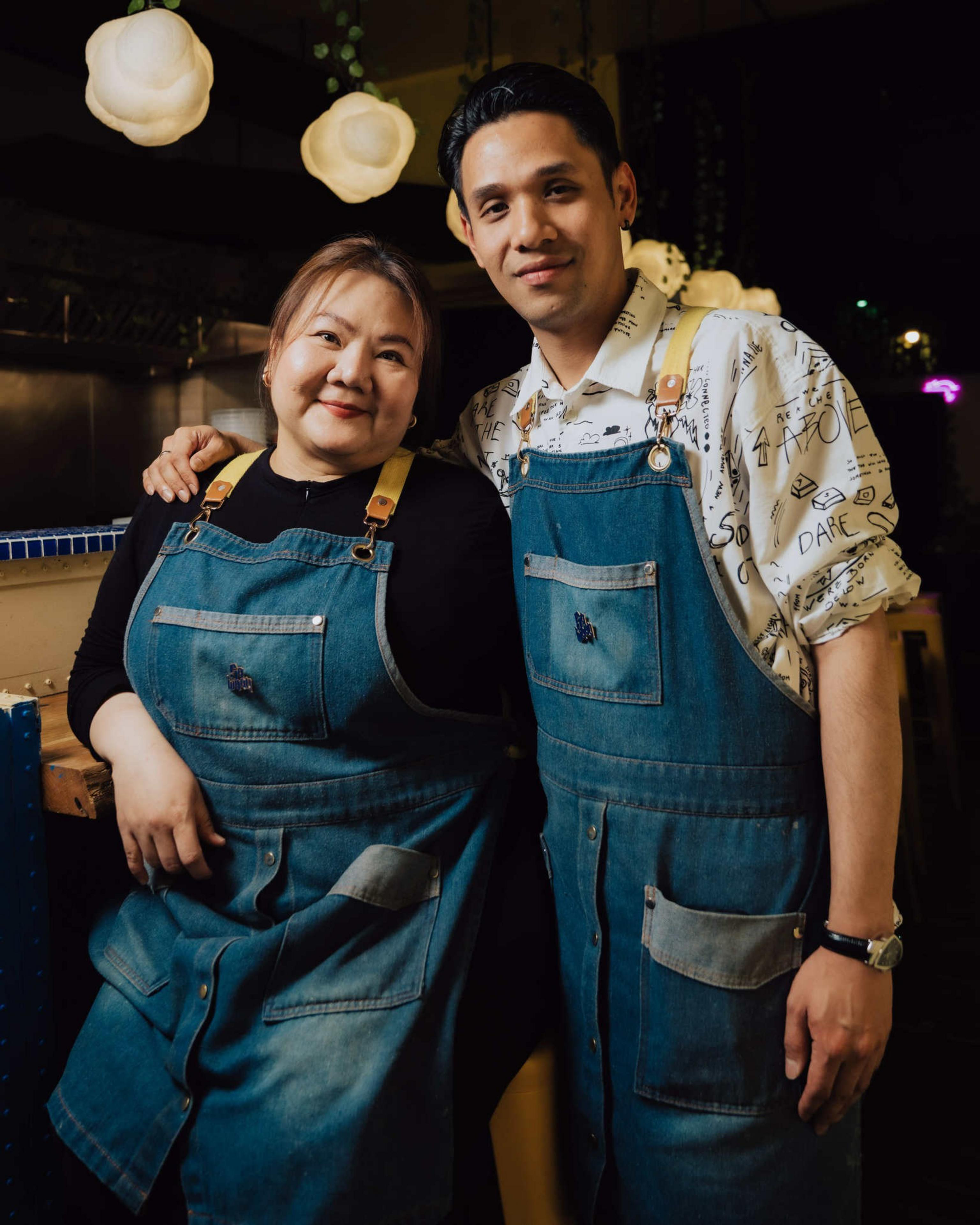 A woman and a man wear matching blue denim aprons, smiling closely together under warm hanging lights in a cozy indoor setting.