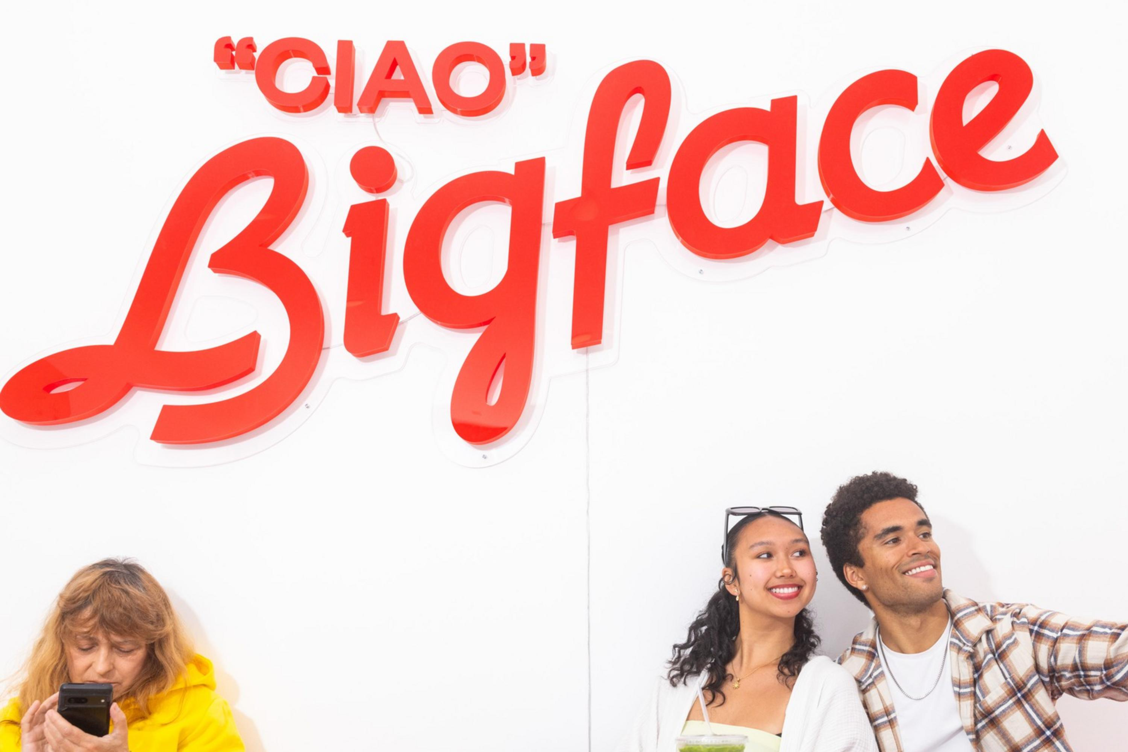 A couple takes a selfie under a red "Ciao Bigface" sign. They are sitting at a small table, and a person in a yellow hoodie is on their phone to the left.