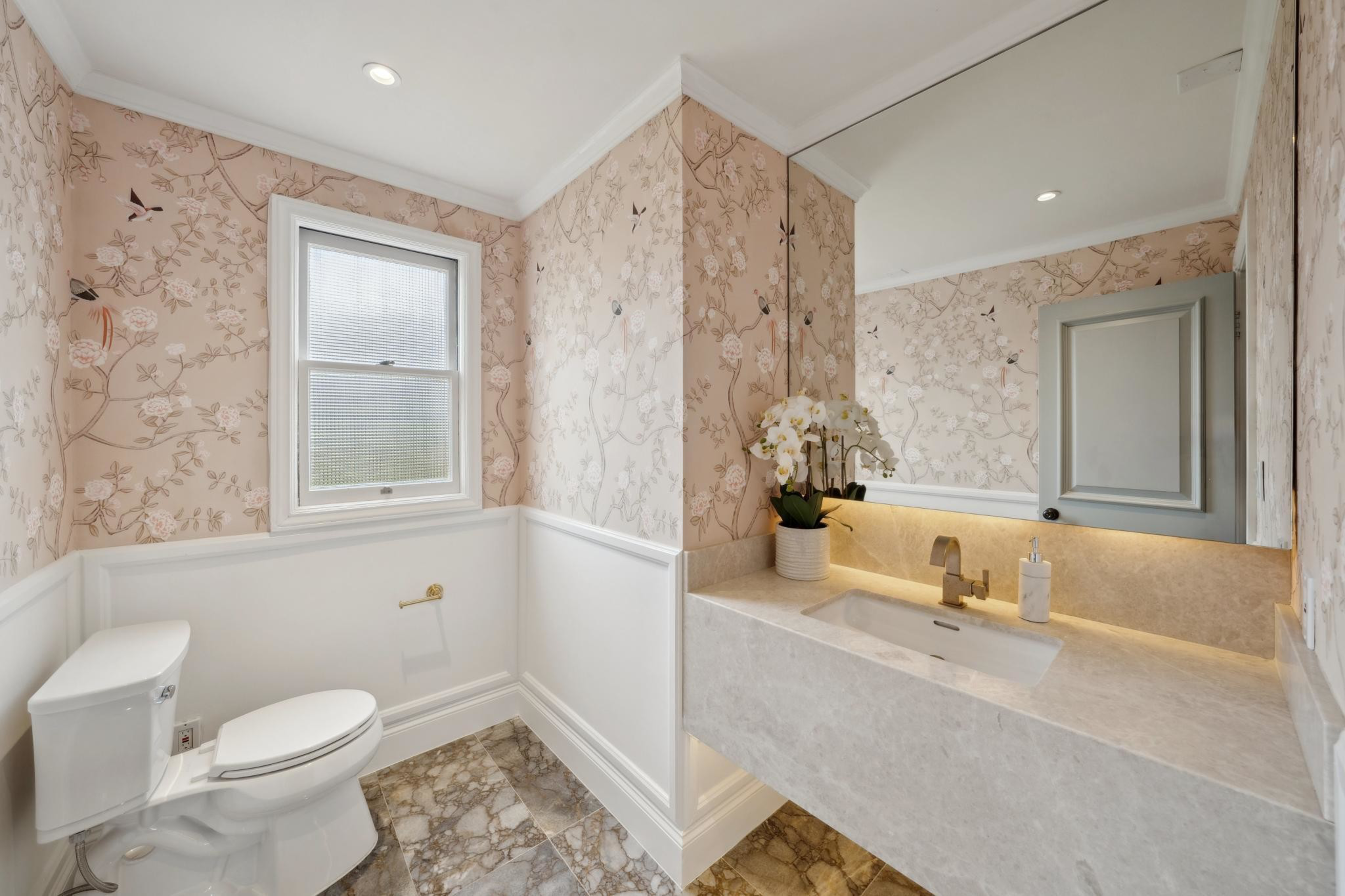 A bathroom with floral wallpaper, a white toilet under a frosted window, a marble floor, and a beige marble sink with a faucet and a potted orchid.
