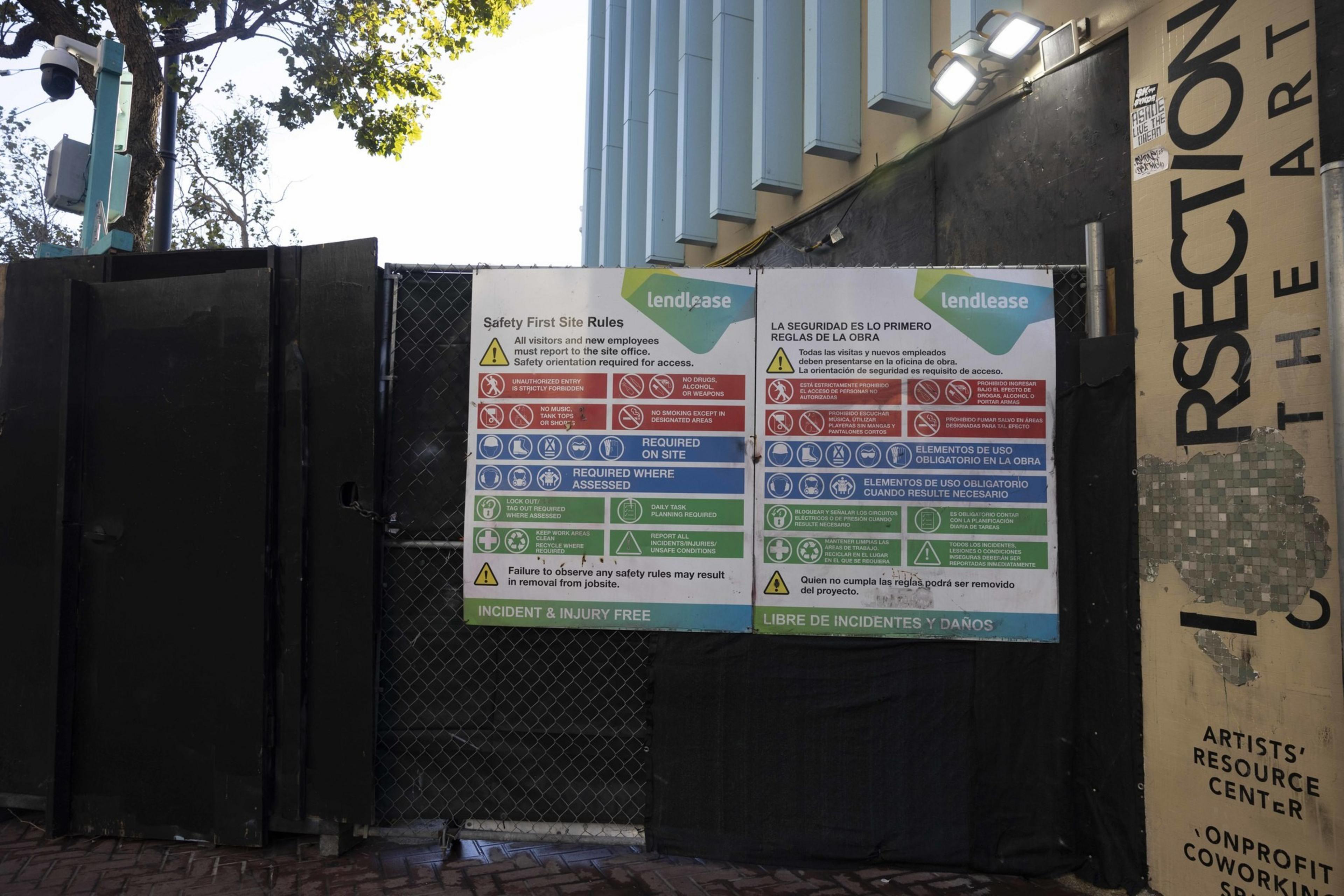 The image shows a construction site entrance with black gates, surveillance cameras, and safety signs in English and Spanish, indicating site rules and required safety measures.