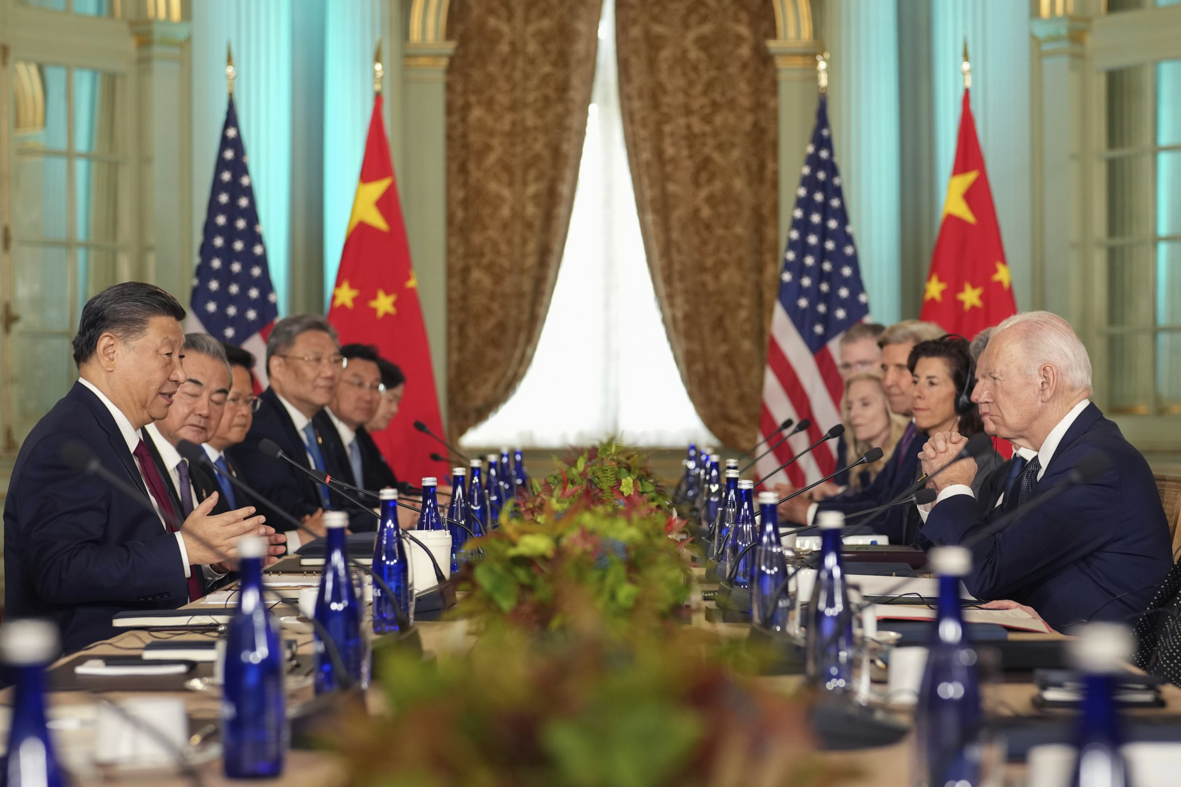President Joe Biden listens as China's President President Xi Jinping speaks while seated at a long table with other delegates listening as well. Two Chinese and two American flags are at the back of the room in a formal dining room setting.