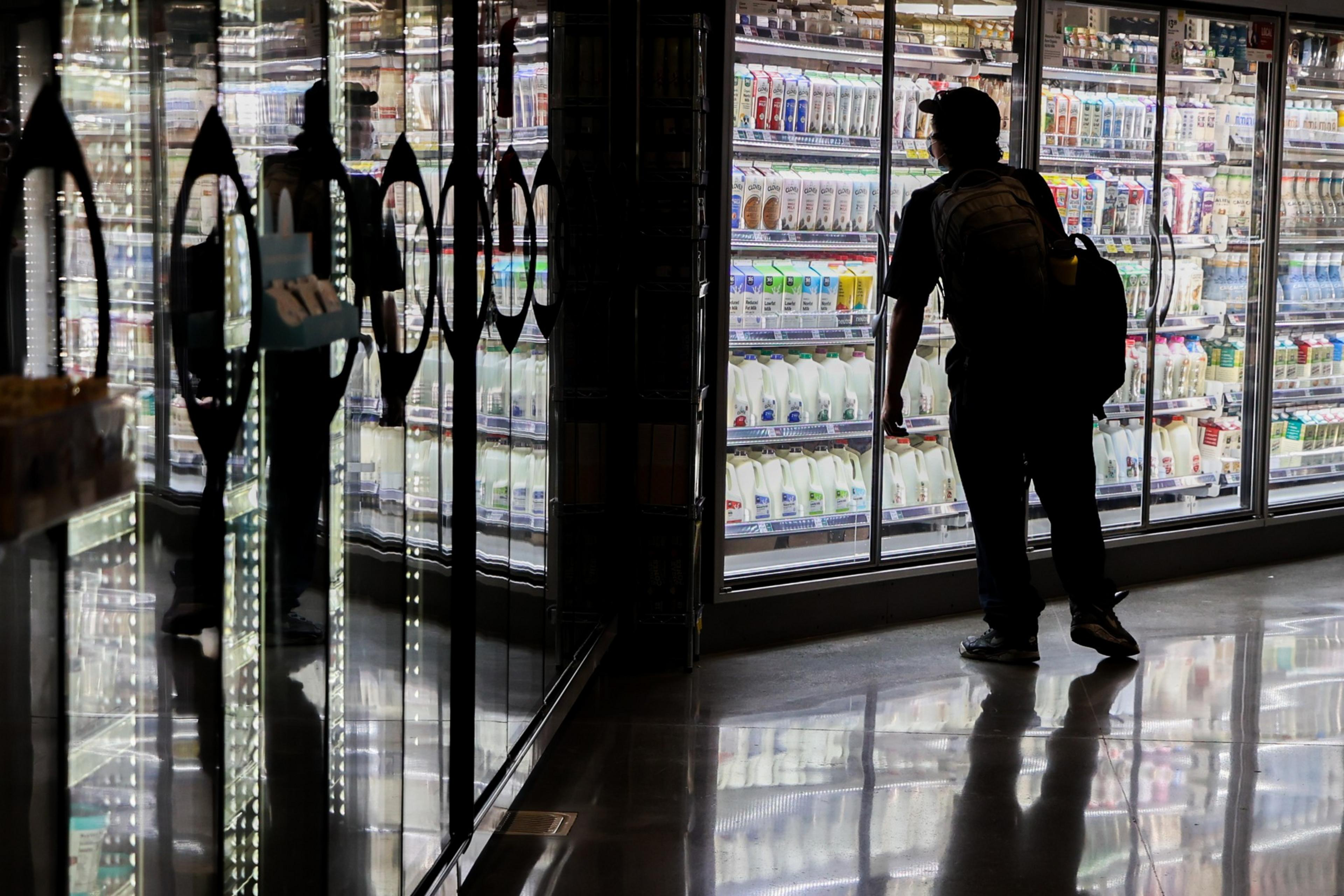 A person wearing a cap and backpack stands in front of brightly lit refrigerated shelves filled with milk and dairy products in a store.