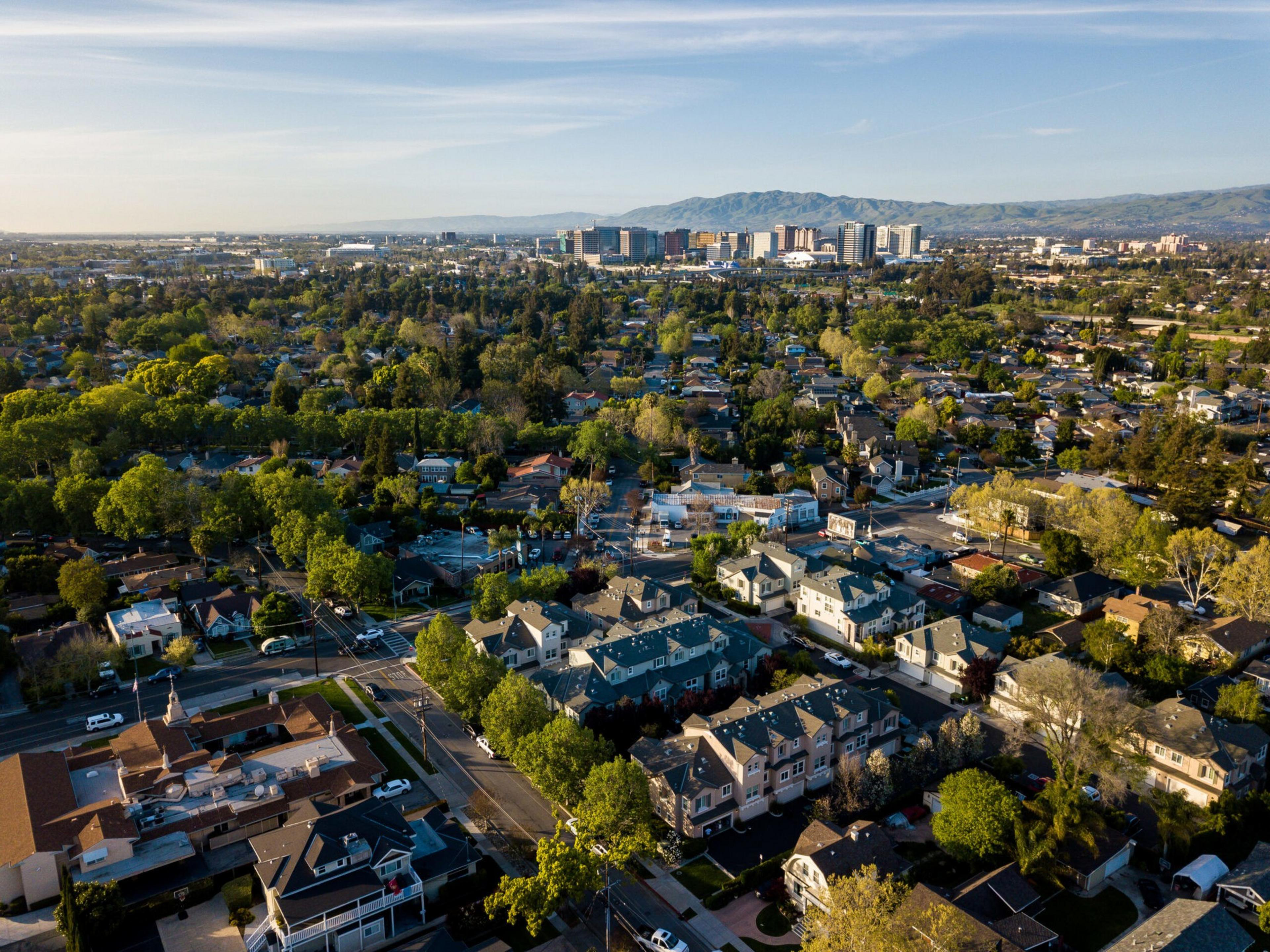 A suburban neighborhood with houses, trees, and streets spreads out toward a distant city skyline and mountain range under a clear sky.
