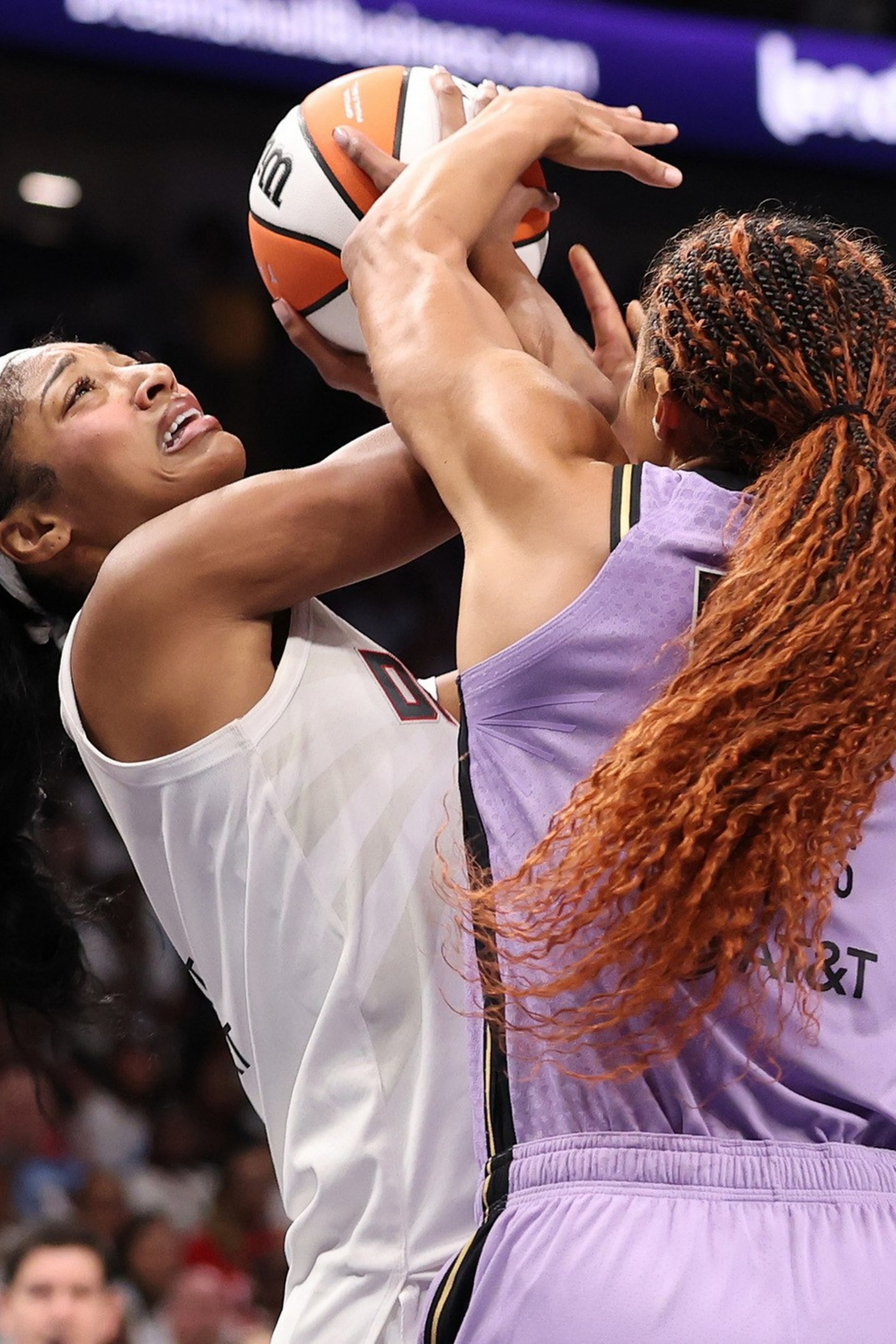 Two basketball players in purple jerseys defend against a player in a white jersey attempting a shot while holding the ball above her head.