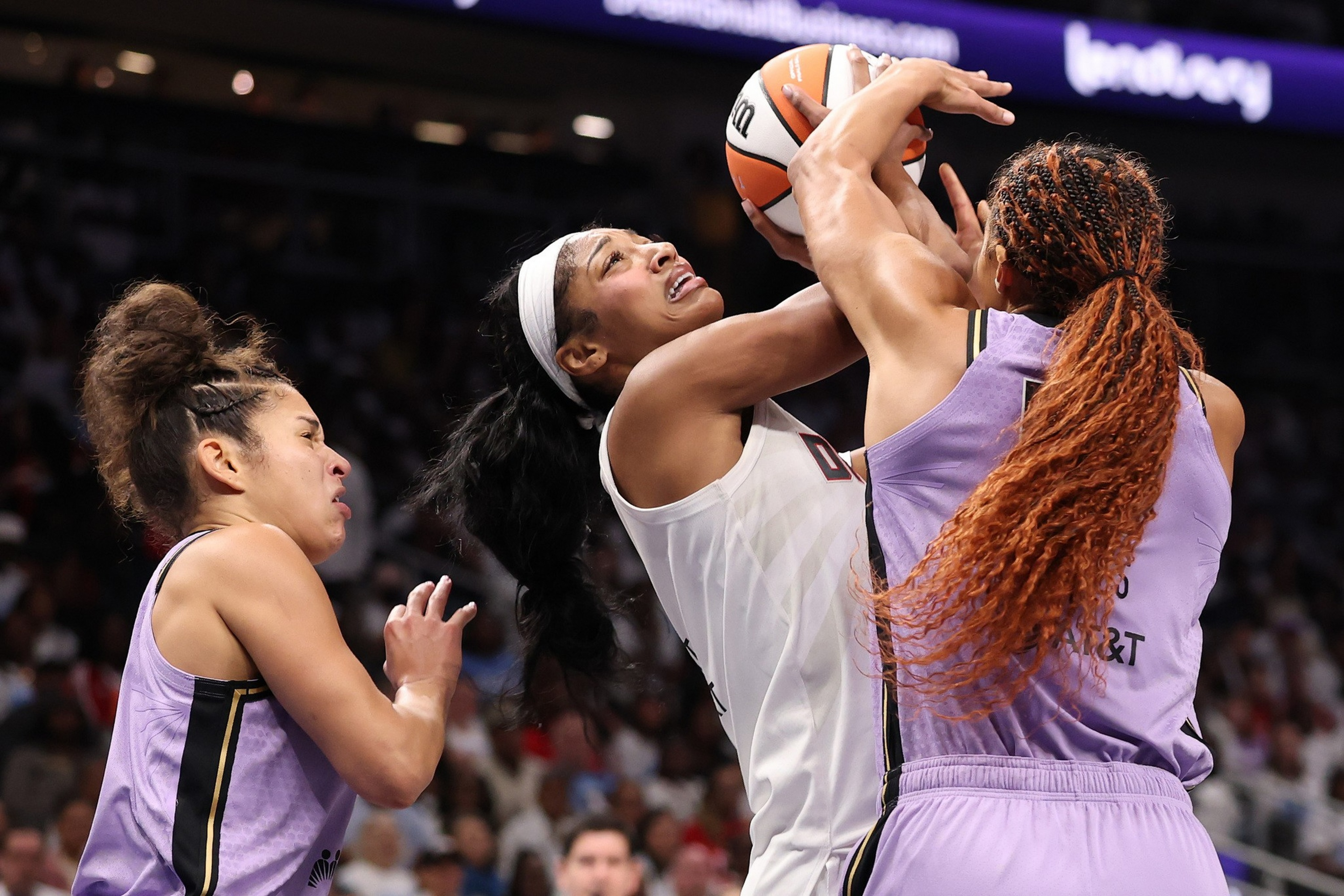 Two basketball players in purple jerseys defend against a player in a white jersey attempting a shot while holding the ball above her head.